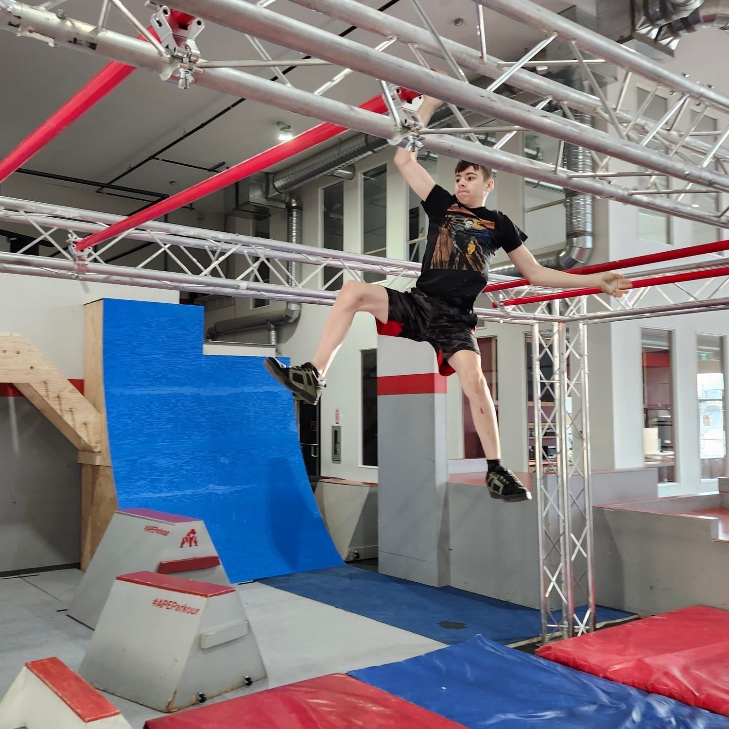 A young man hangs from an overhead bar on an indoor obstacle course.