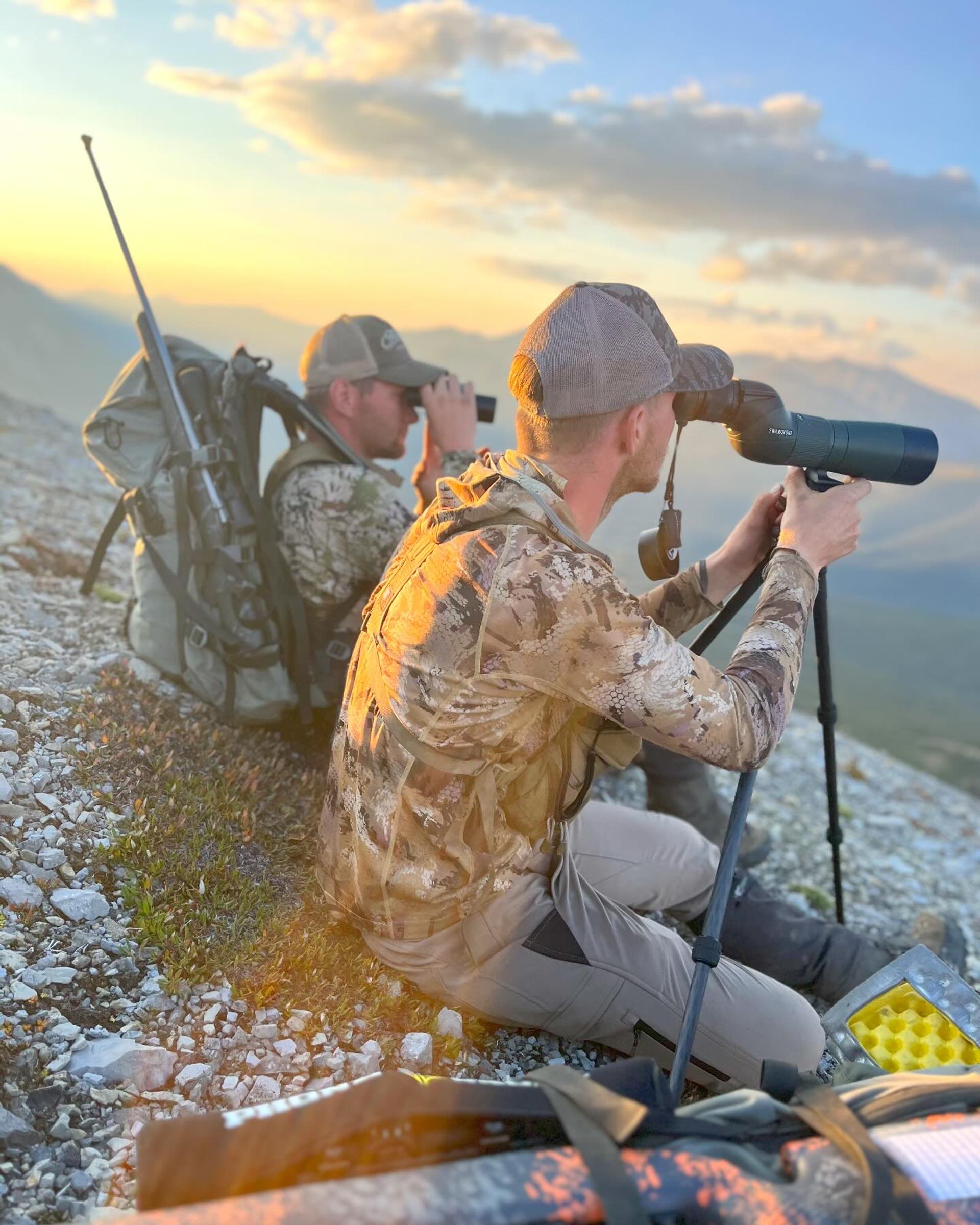 Two people in camo gear using binoculars and scope on rocky terrain at sunrise or sunset.