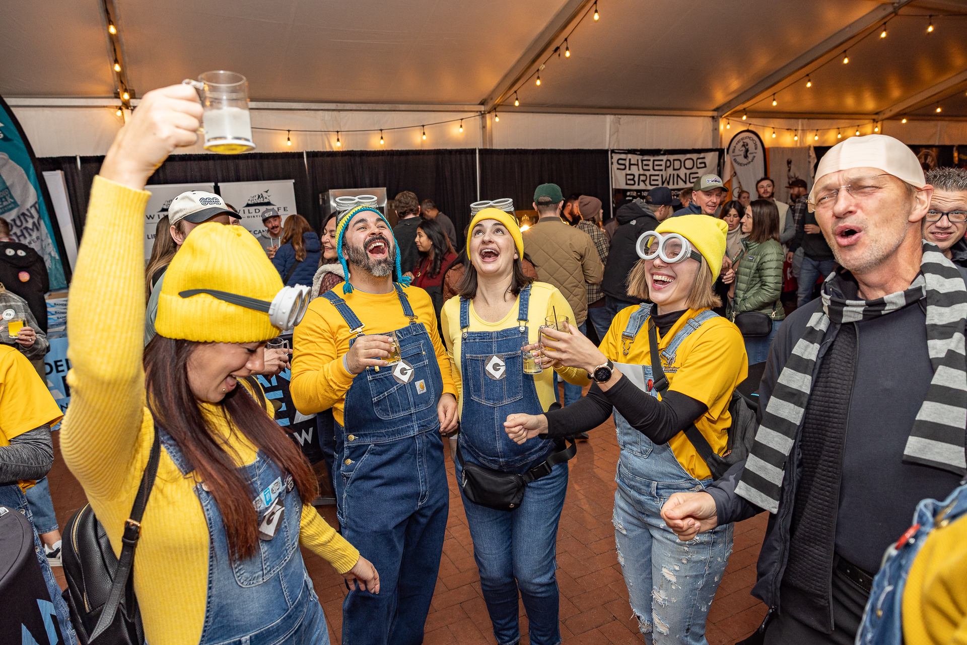 Adults in Minion costumes hold drinks and sing together at a crowded indoor event.