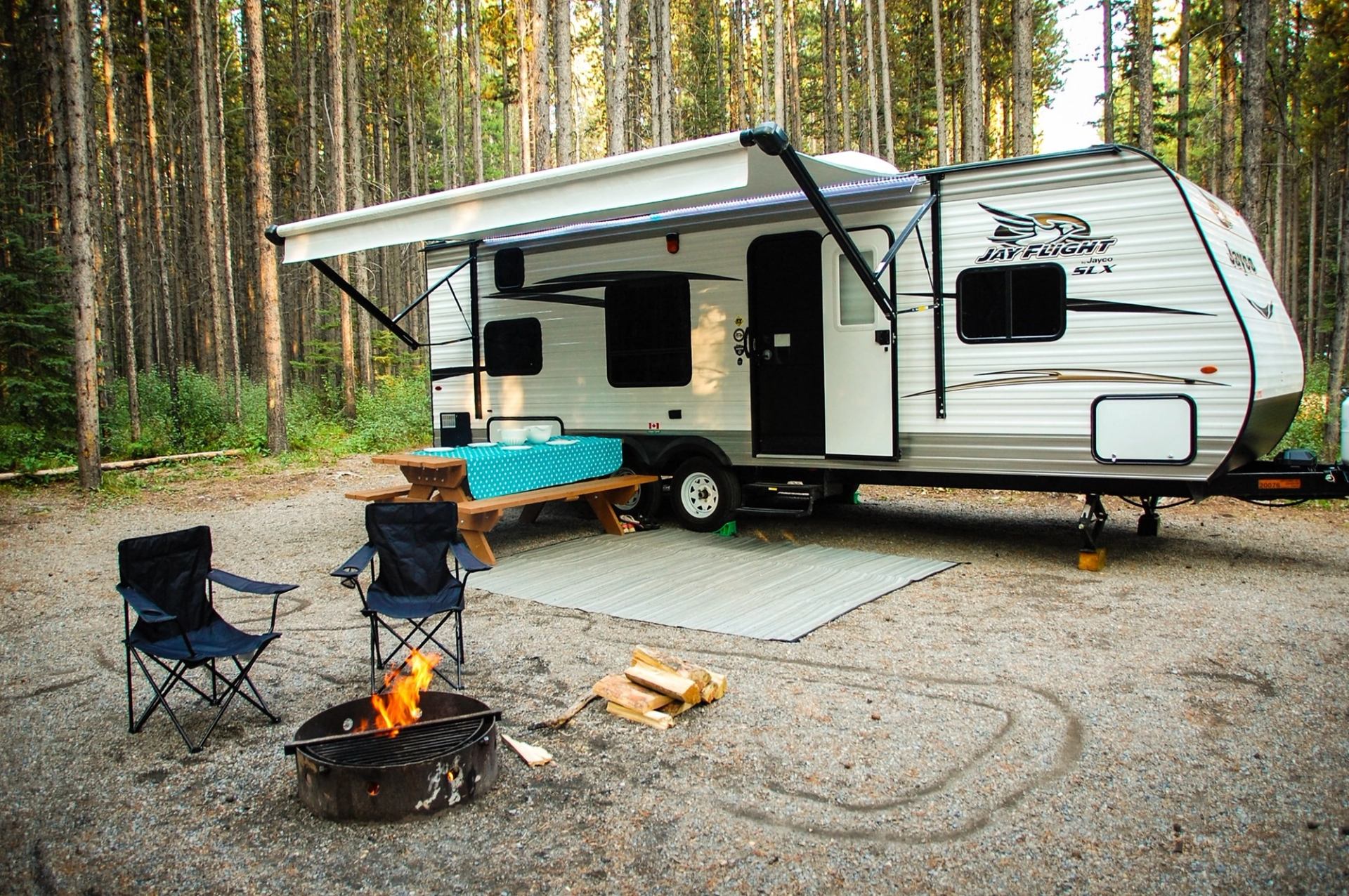 Travel trailer with awning, campfire, and picnic setup in forested campsite.