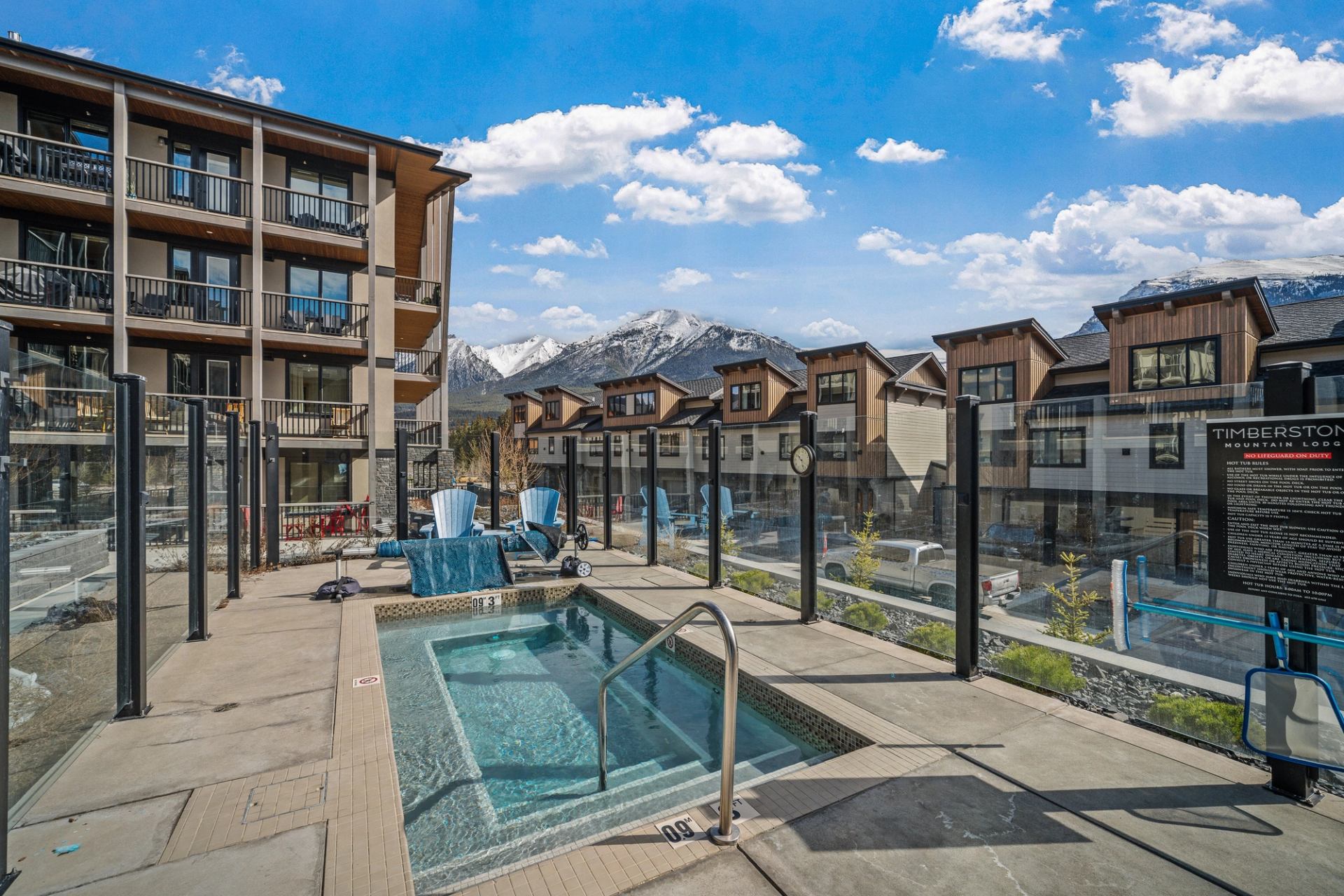 Outdoor hot tub with glass fence, lounge chairs and mountain view behind nearby buildings.