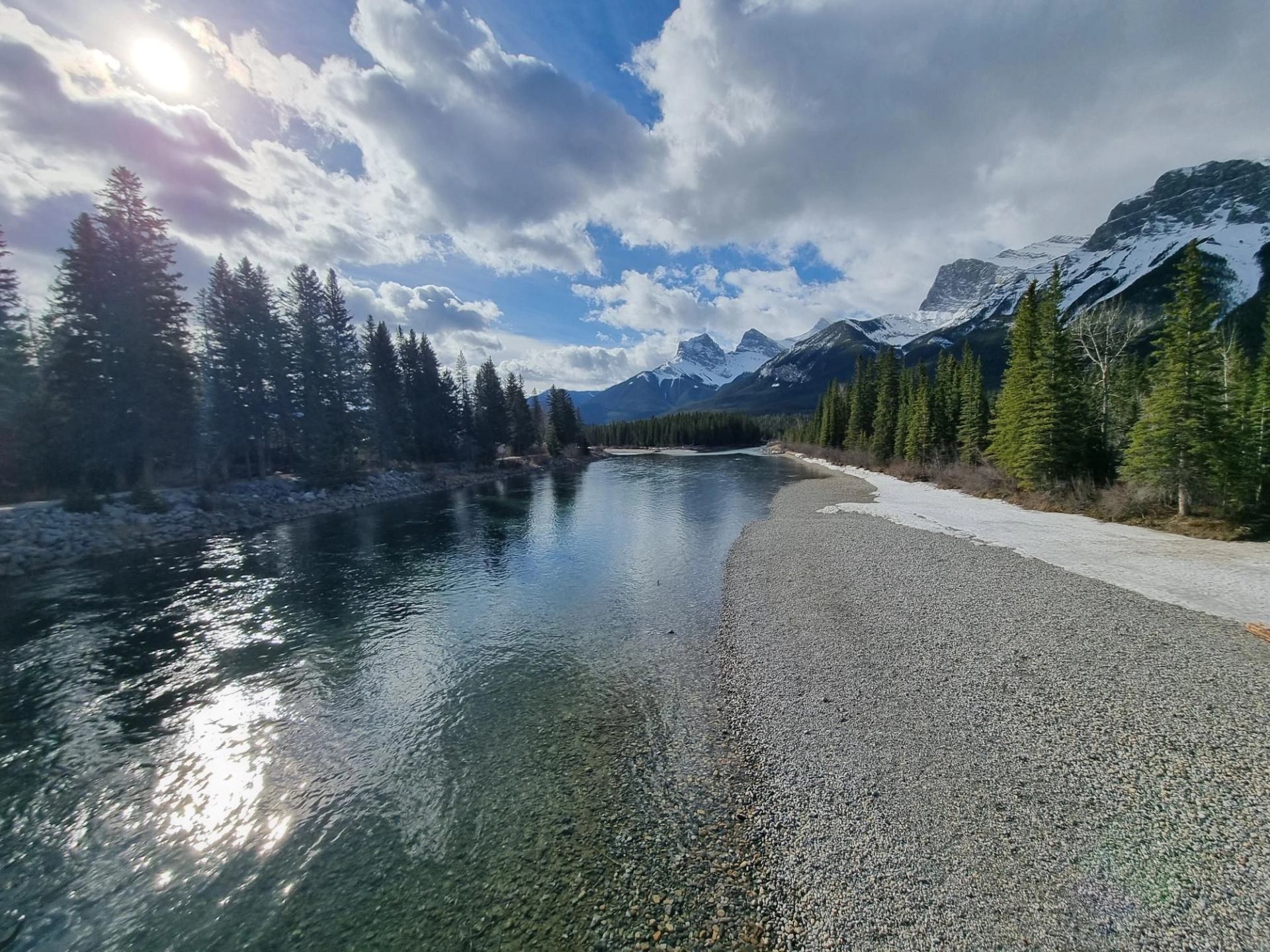 A calm river curving past evergreens and mountains under a partly cloudy sky at sunset.