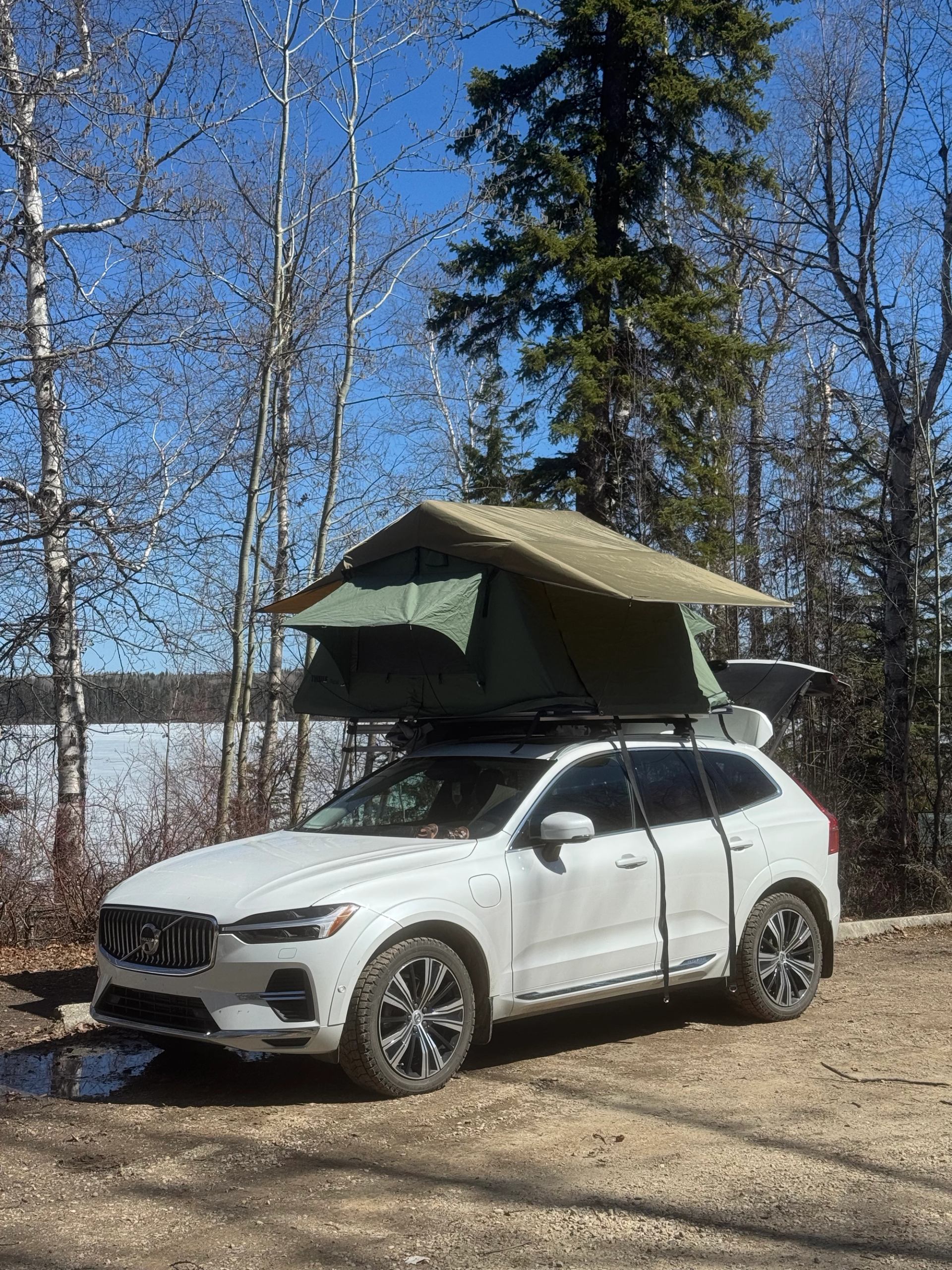 White SUV with rooftop tent parked on dirt road beside frozen lake and trees.