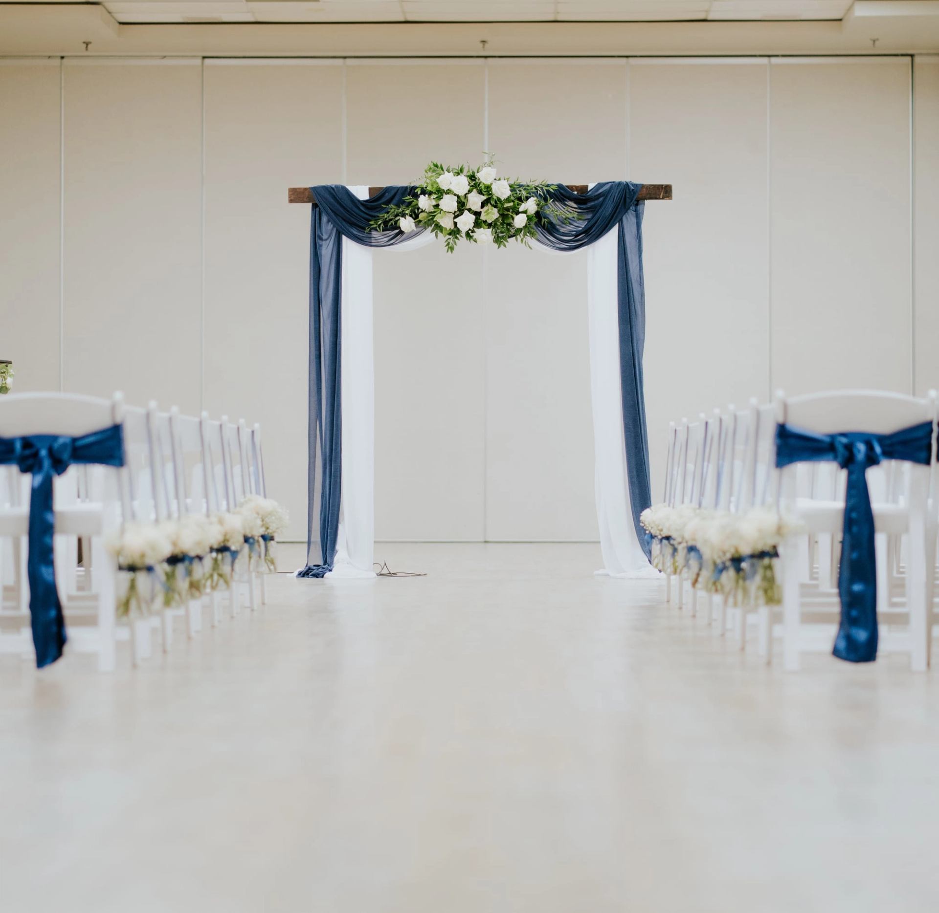 Ceremony aisle with white chairs, blue bows and a flower-draped arch.