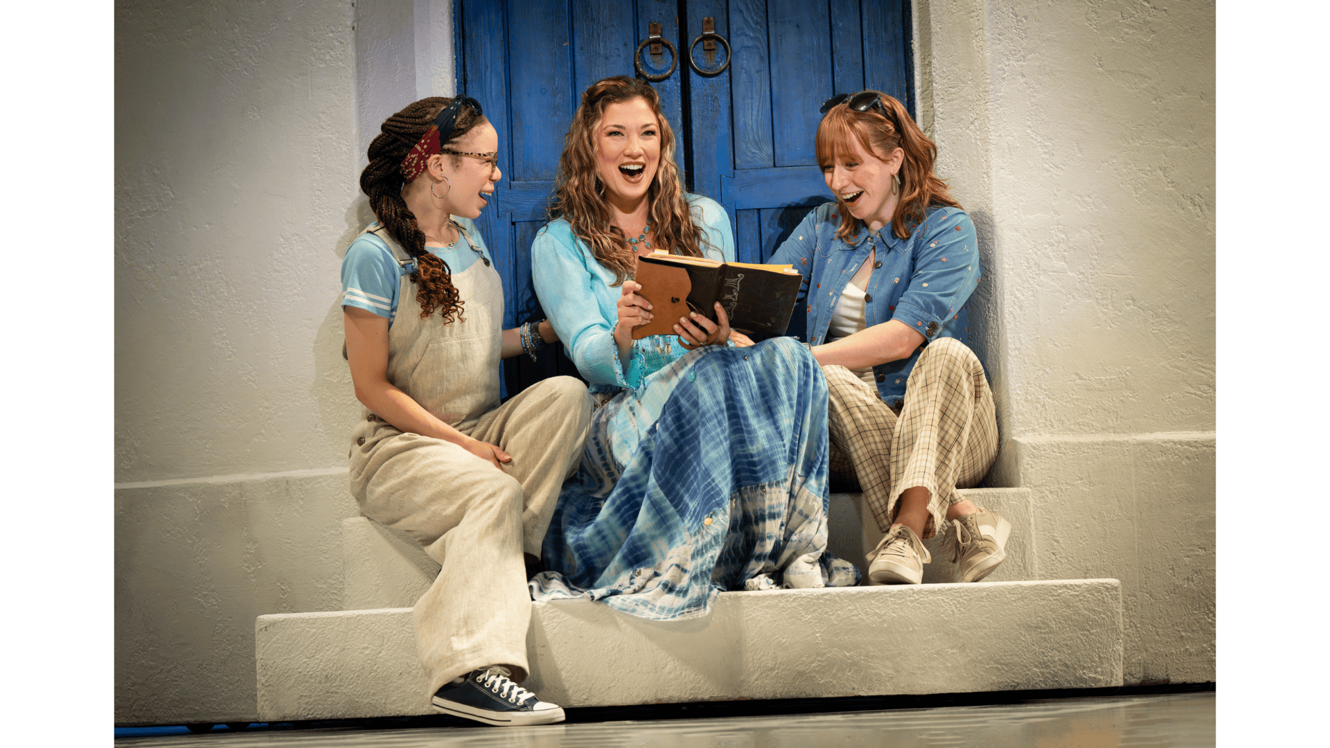 Three women sit in a theatre box holding scripts in front of blue doors.