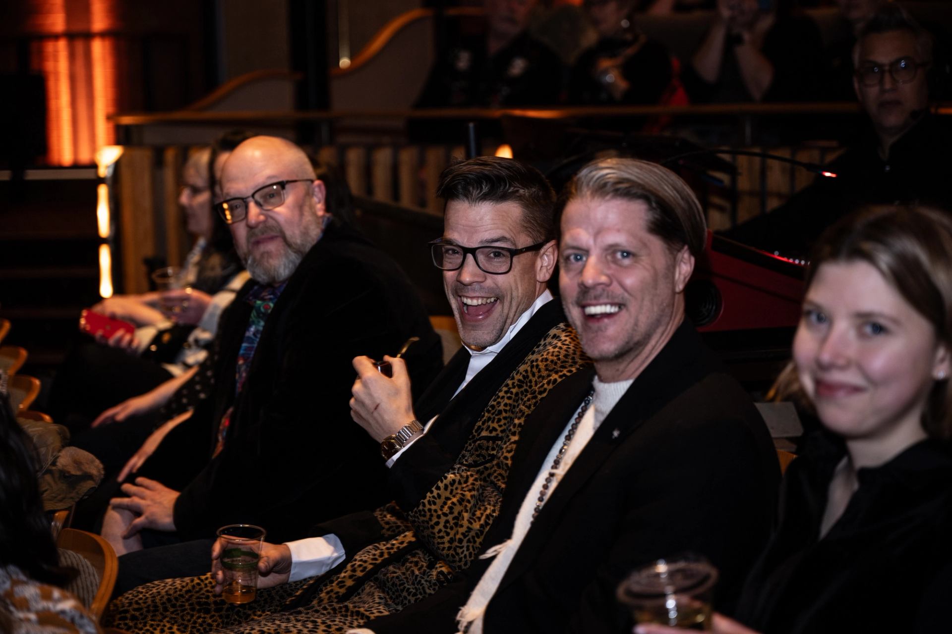 Two men laugh, one wearing a leopard print jacket, and a woman smiles in a dimly lit audience.