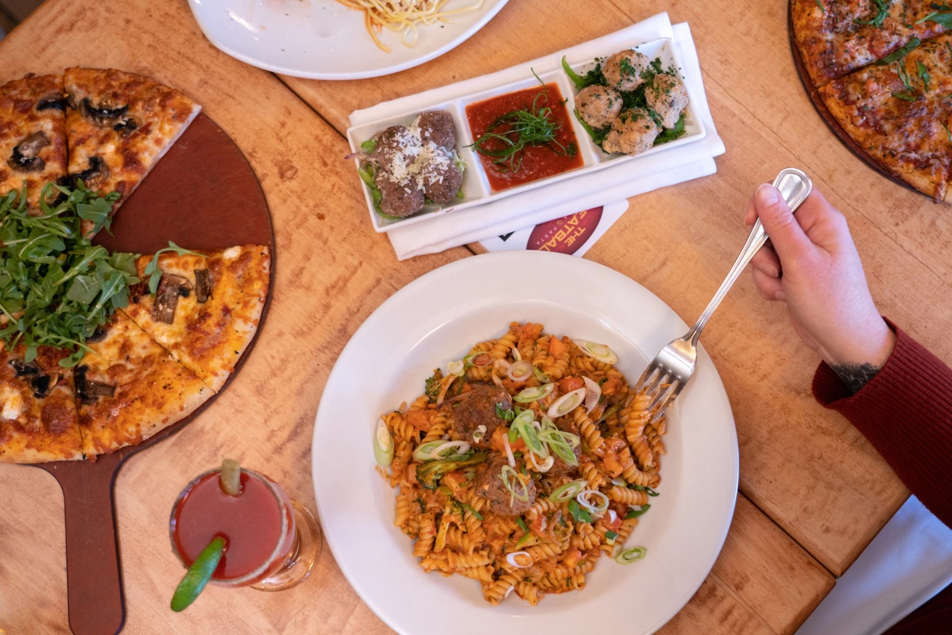 Table with pizza, pasta, meatballs, sauces, and a lime-garnished drink.