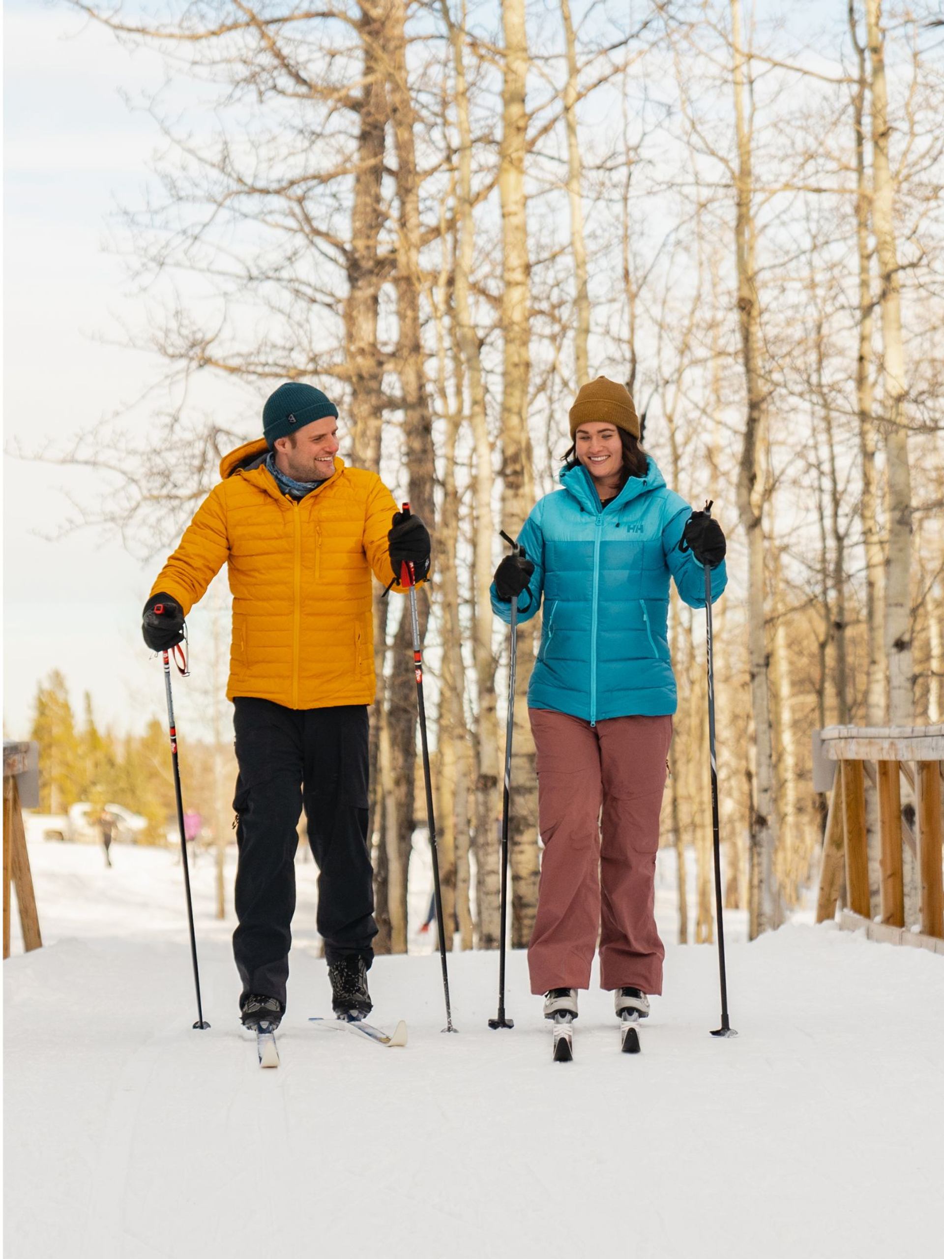 Skiers in winter jackets stand on snow with poles in a wooded path.