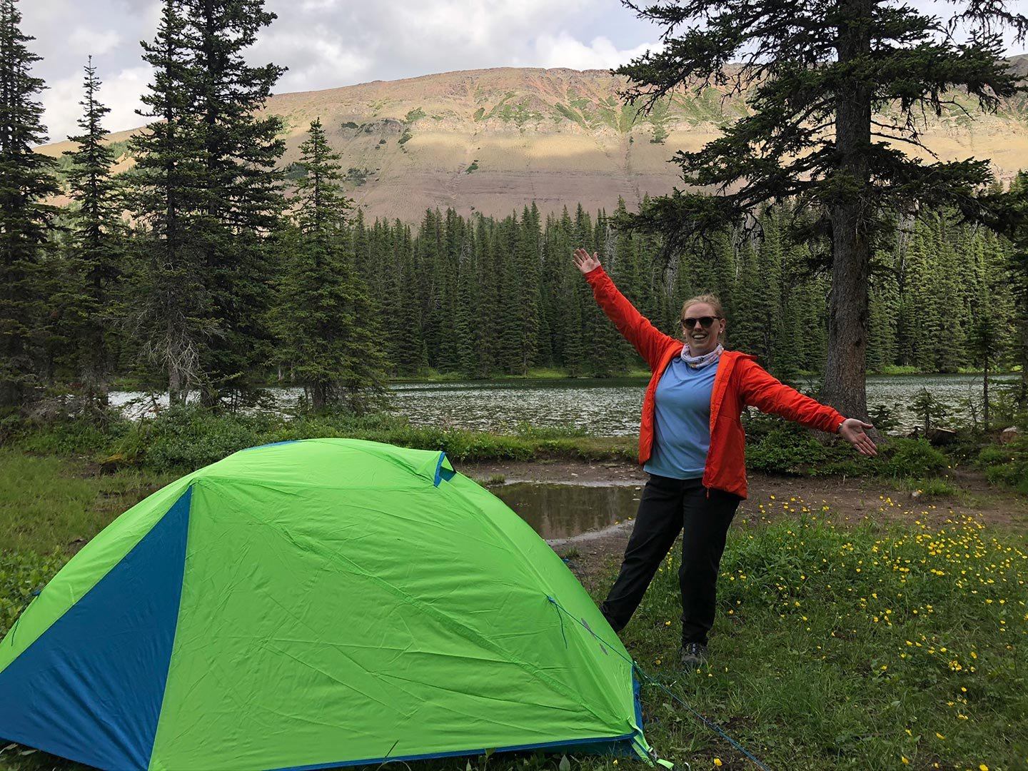 Camper standing beside bright green tent near forested lake and distant rocky ridge.