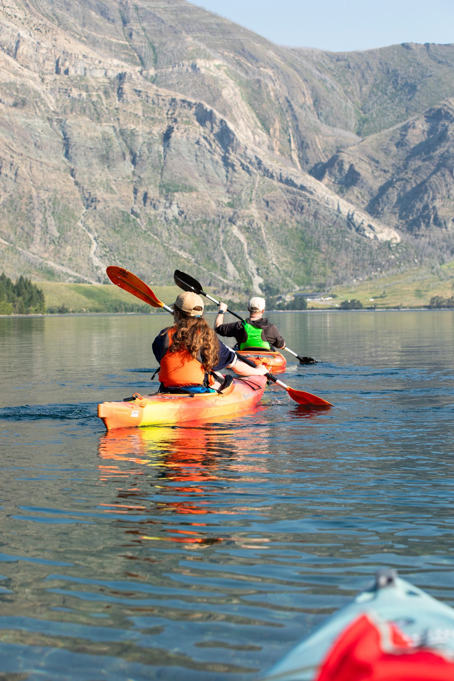 Kayakers paddle across calm lake water with steep rocky mountains in the background.