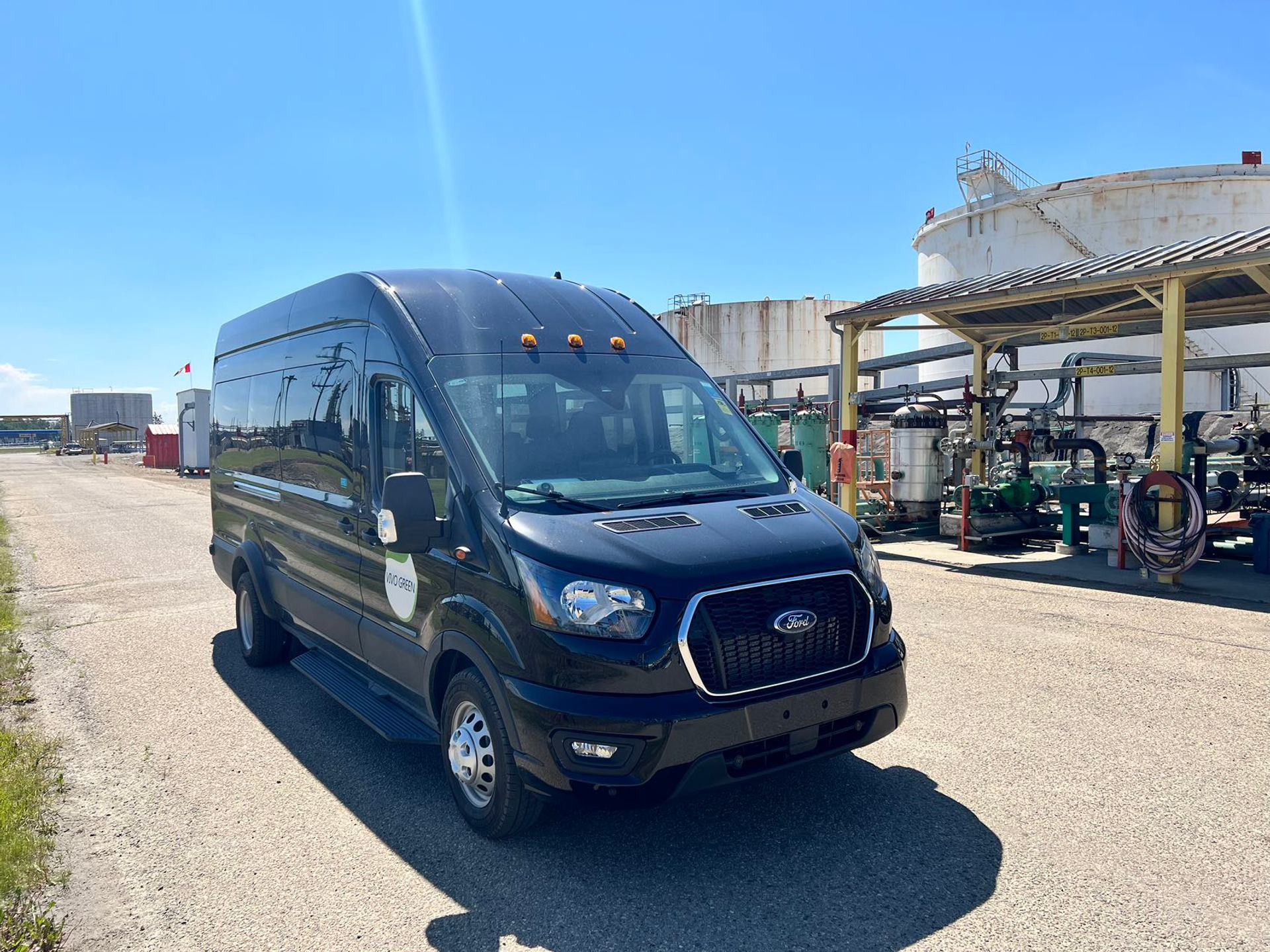 Black Ford cargo van parked beside storage tanks and industrial piping.