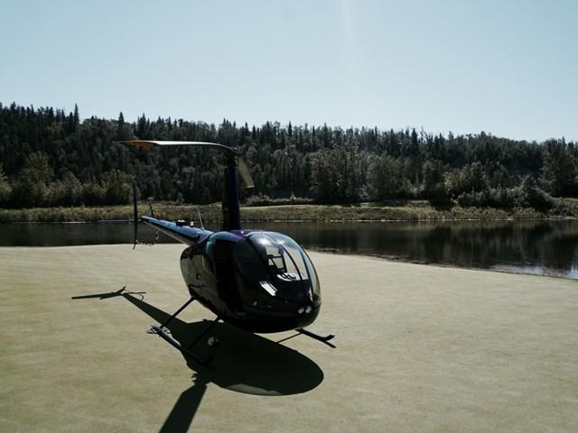 Helicopter parked on a gravel pad beside a lake, with forested hills in background.