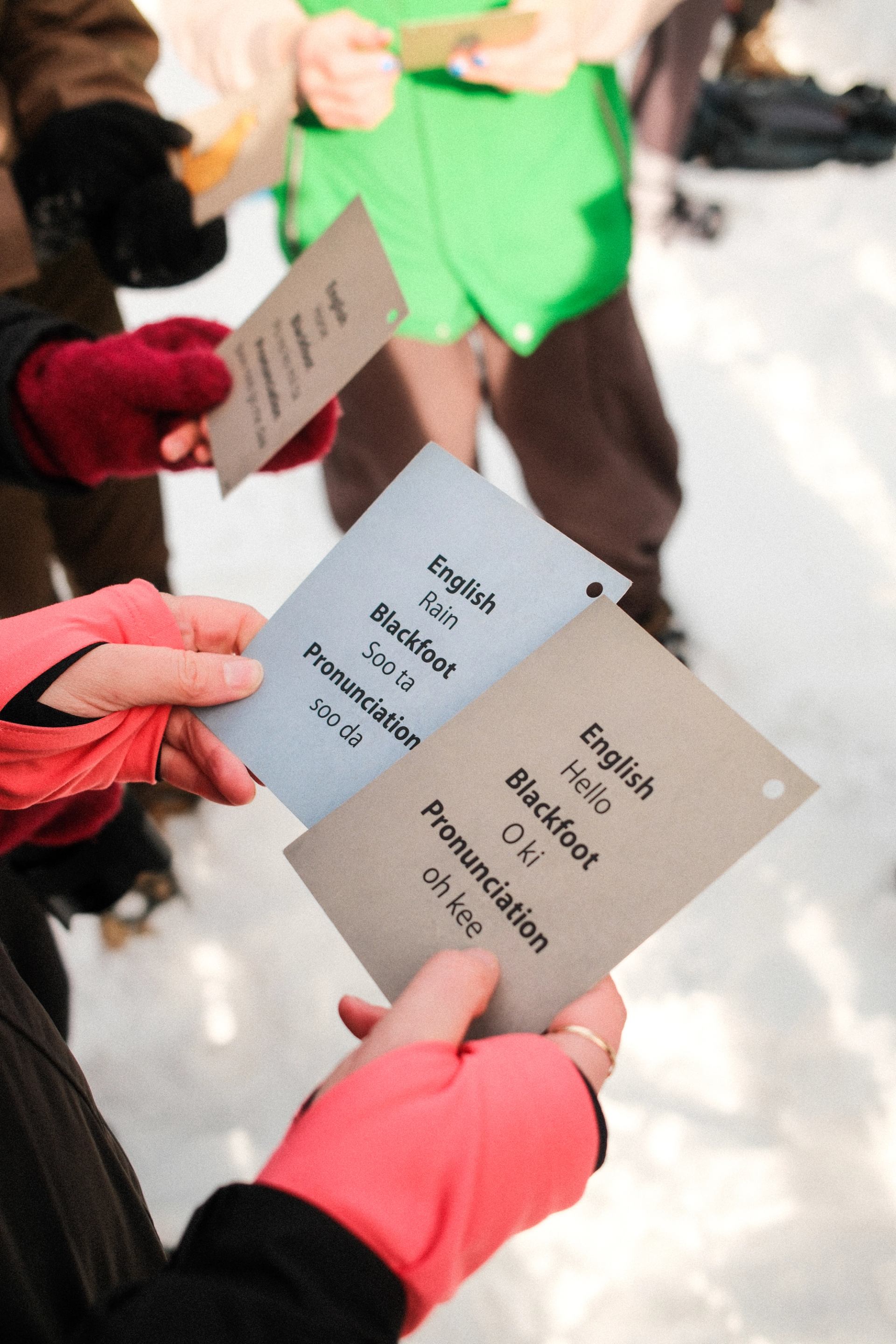 Gloved hands hold language cards showing English words, Blackfoot translations and pronunciations.