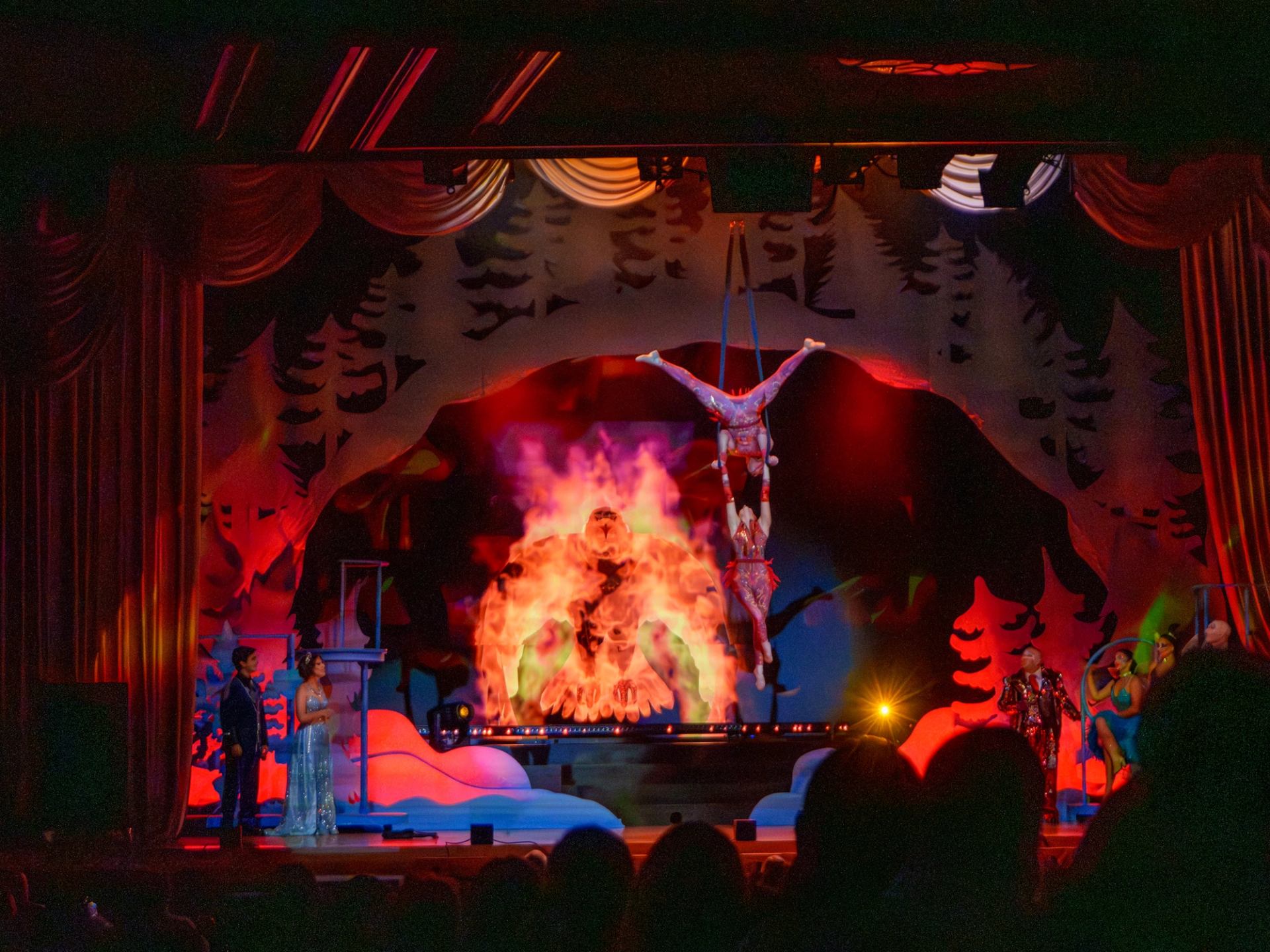 Acrobats hang above a fiery stage projection as performers watch from both sides.