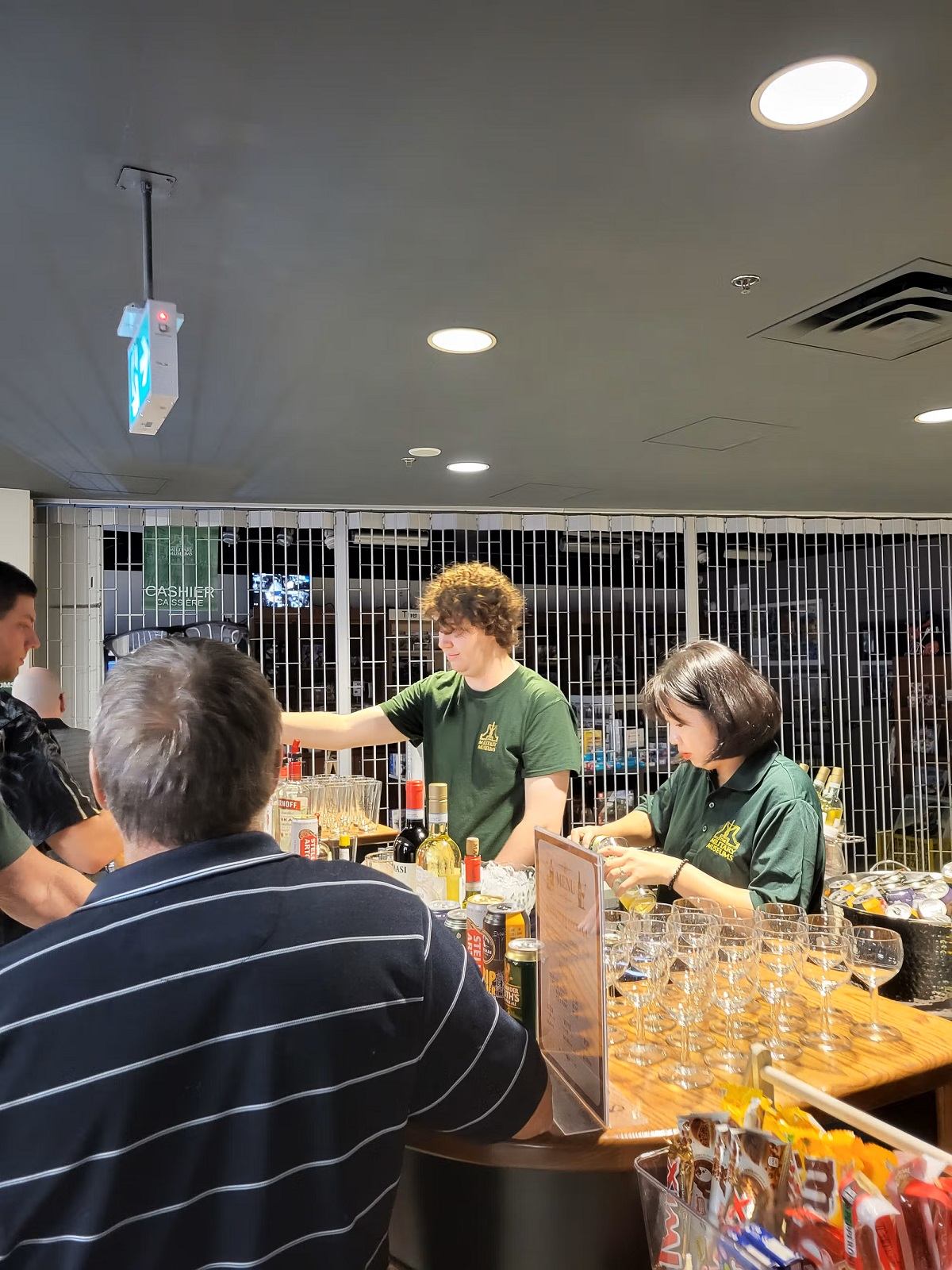 Bartenders in green shirts serve drinks to customers at a bar with bottles and glasses.