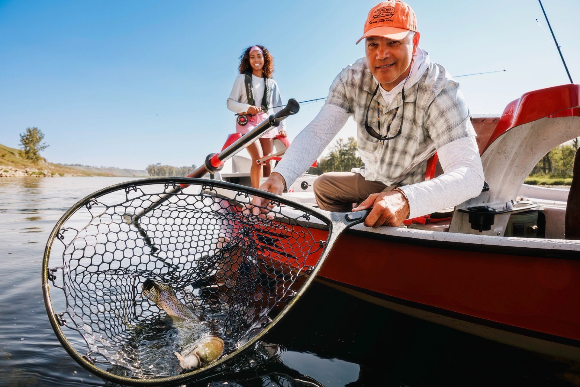 A man in a boat holds a net with a fish in the water, while a woman smiles in the background.