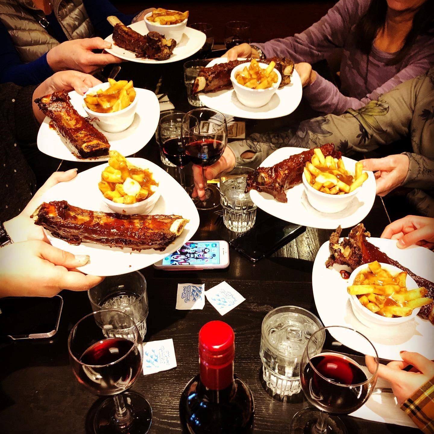 Group toasting with plates of ribs and poutine, surrounded by wine, water glasses, and a wine bottle.