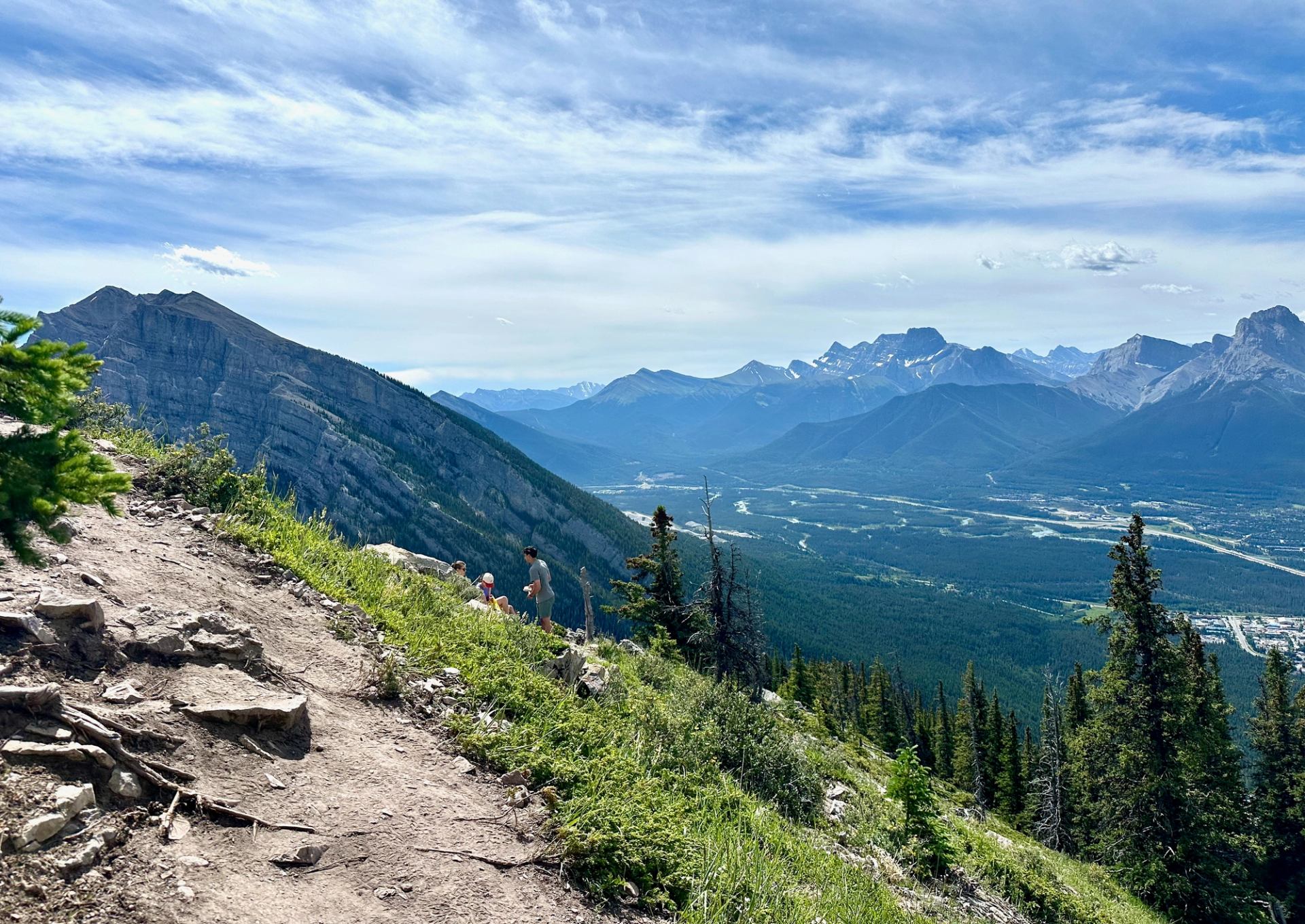 Rocky mountain hiking trail overlooking forested valley, river and distant jagged peaks.