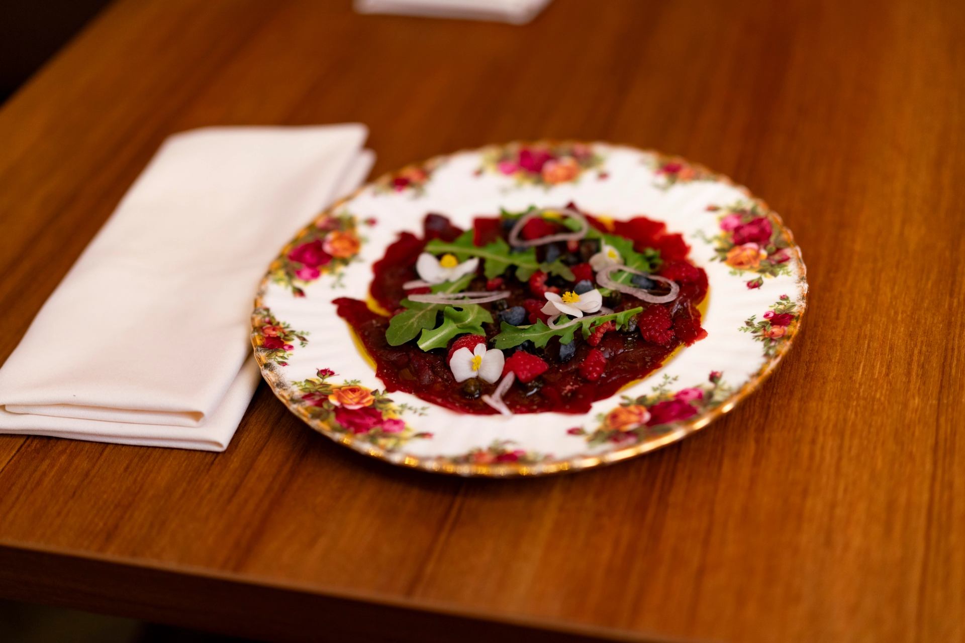 Floral china plate with beet carpaccio, berries, greens and edible flowers on table.