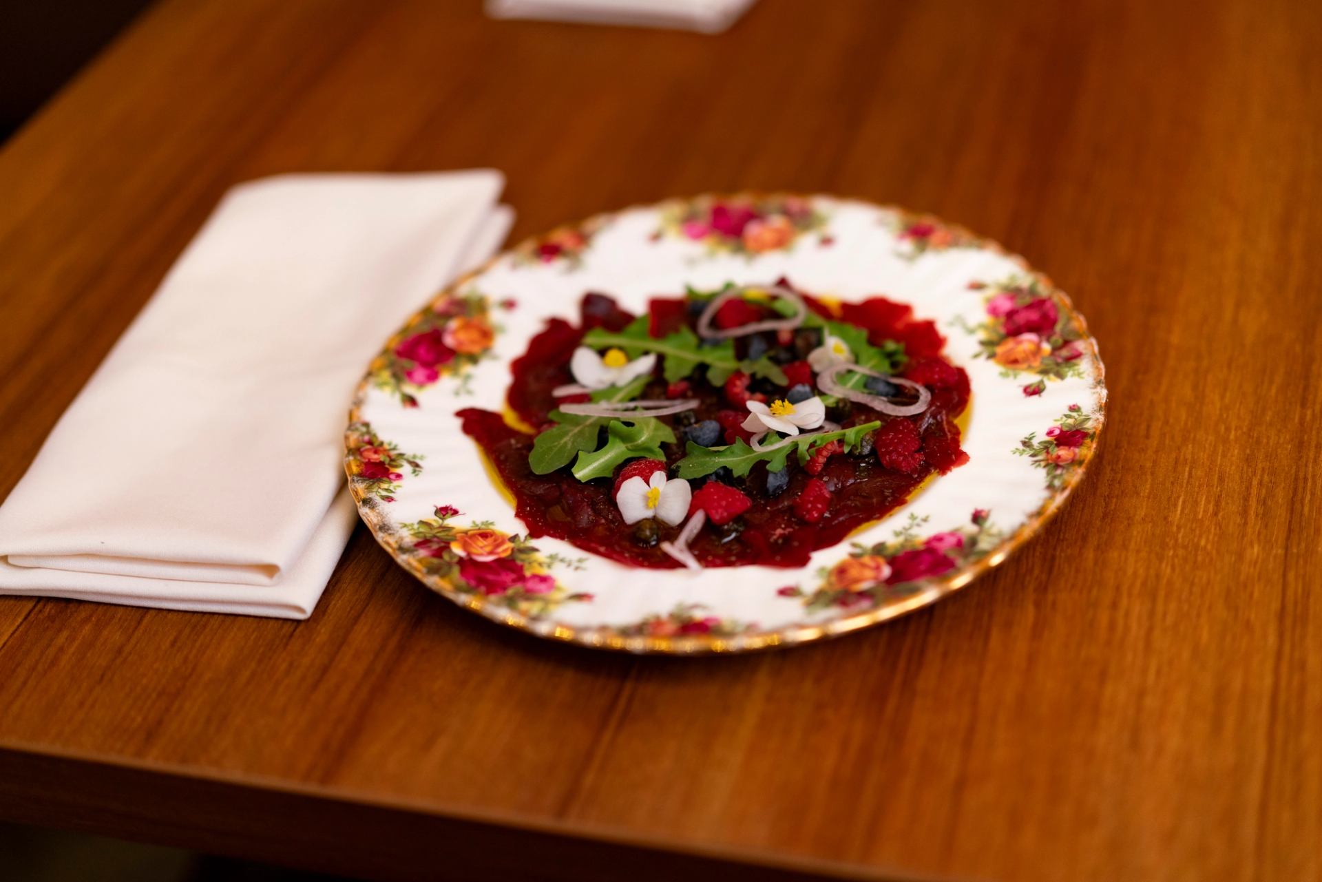Floral china plate with beet carpaccio, berries, greens and edible flowers on table.