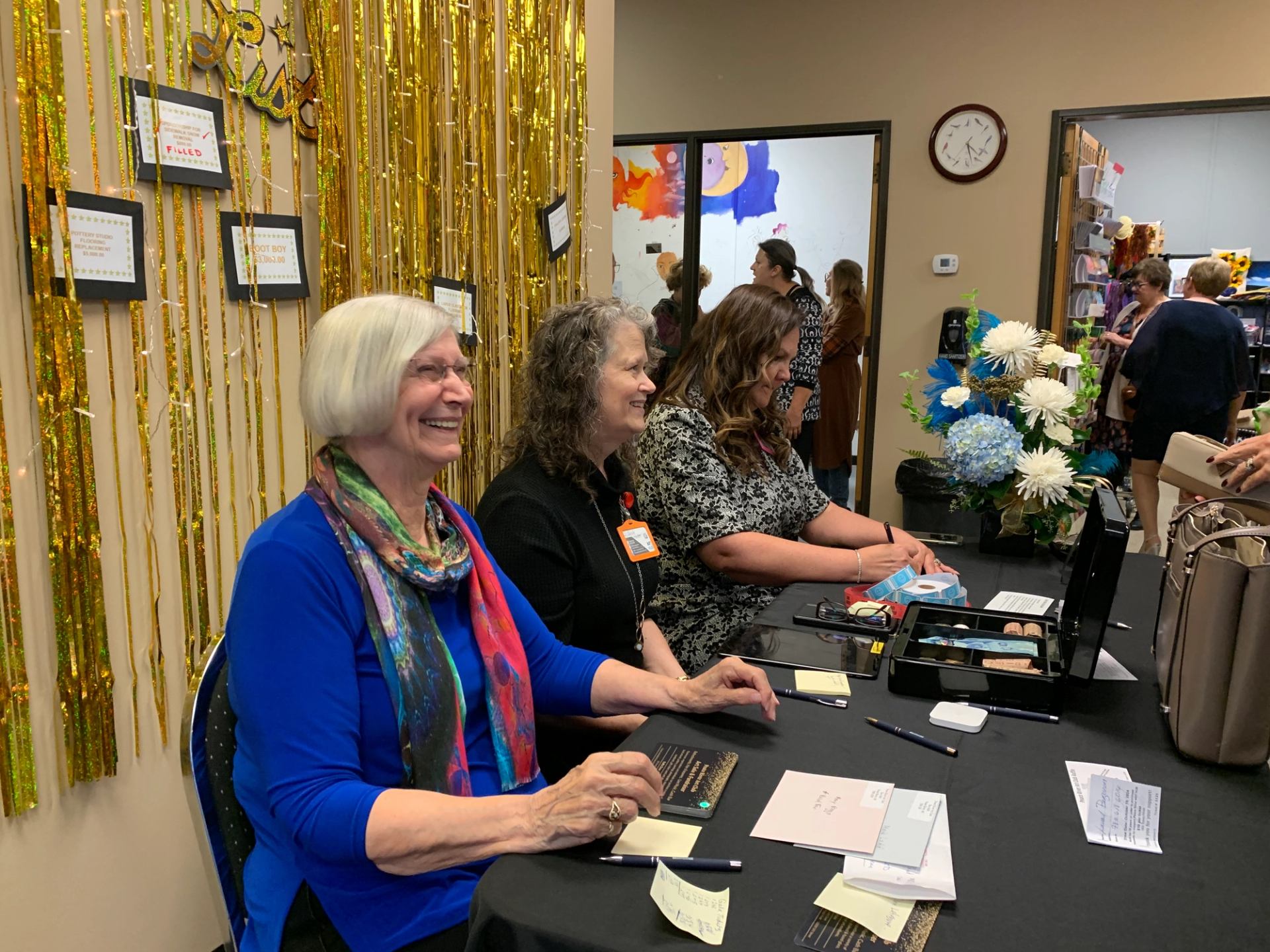 Three people at check-in table with gold streamers and framed awards in background.