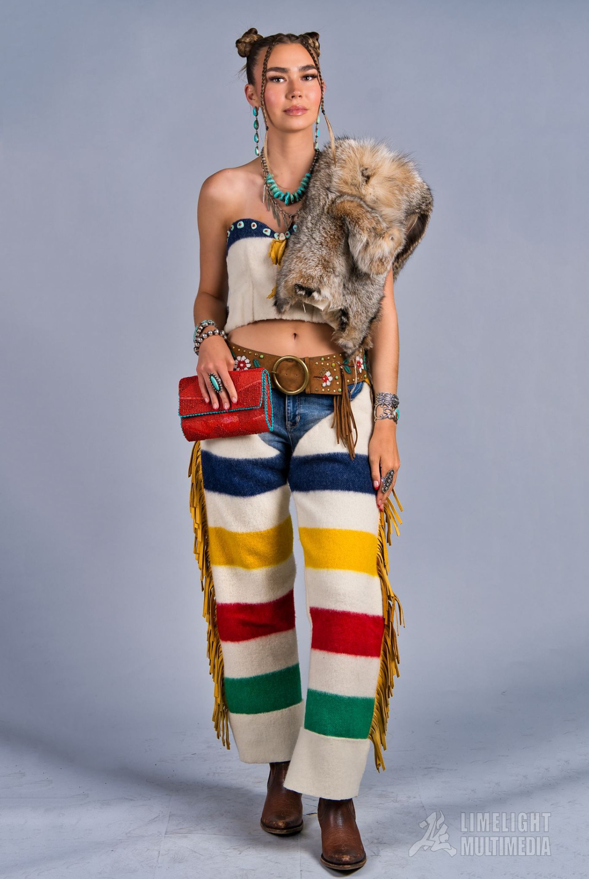 Person in colourful striped pants, fur pelt shawl and beaded accessories against plain backdrop.