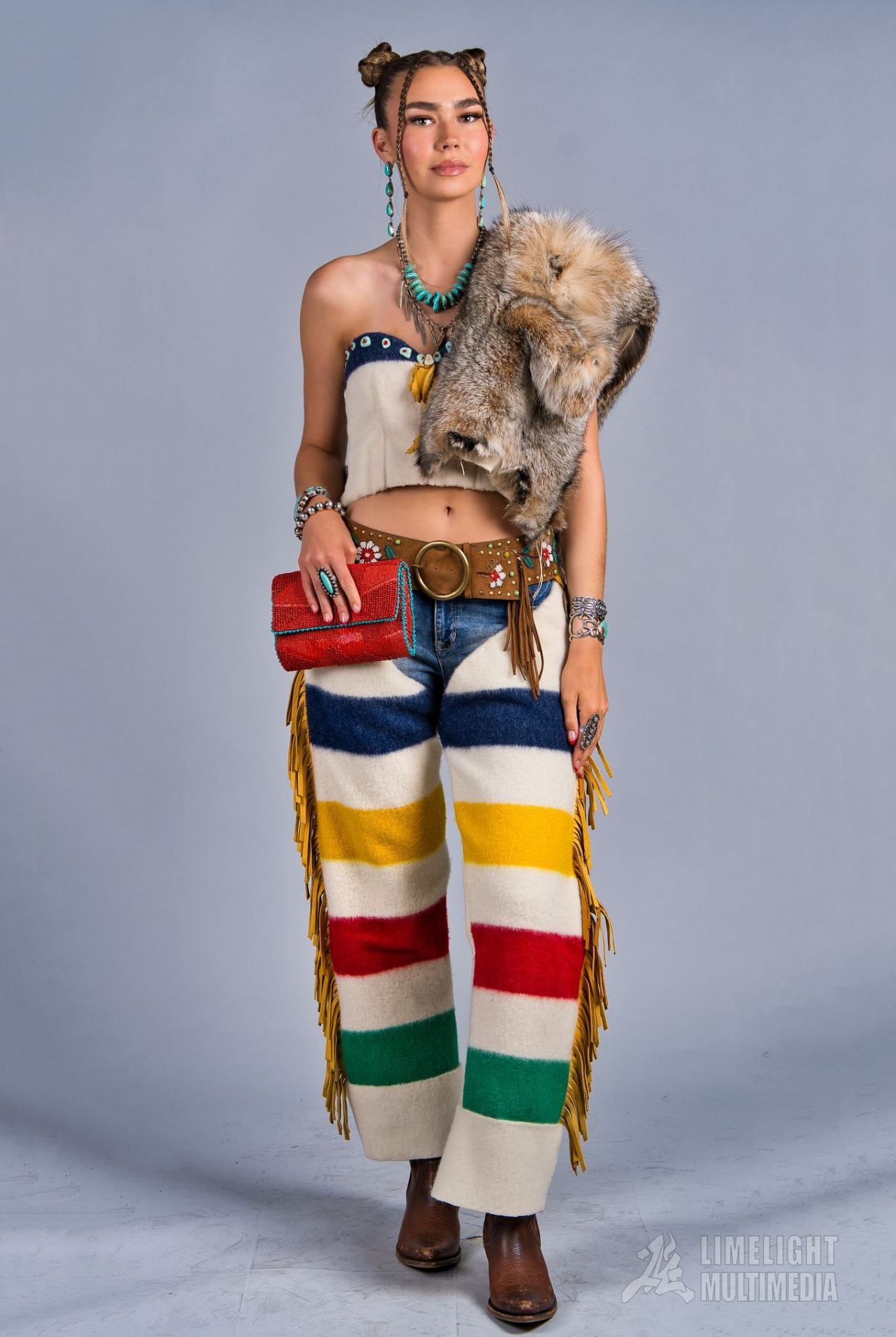 Person in colourful striped pants, fur pelt shawl and beaded accessories against plain backdrop.