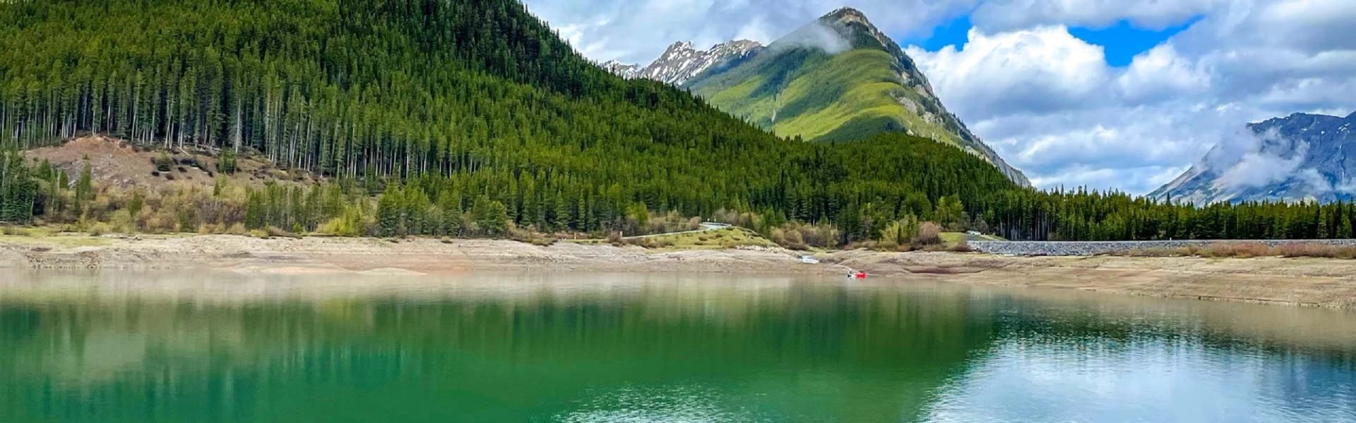 Green lake reflecting forested mountains under partly cloudy sky with low mist.