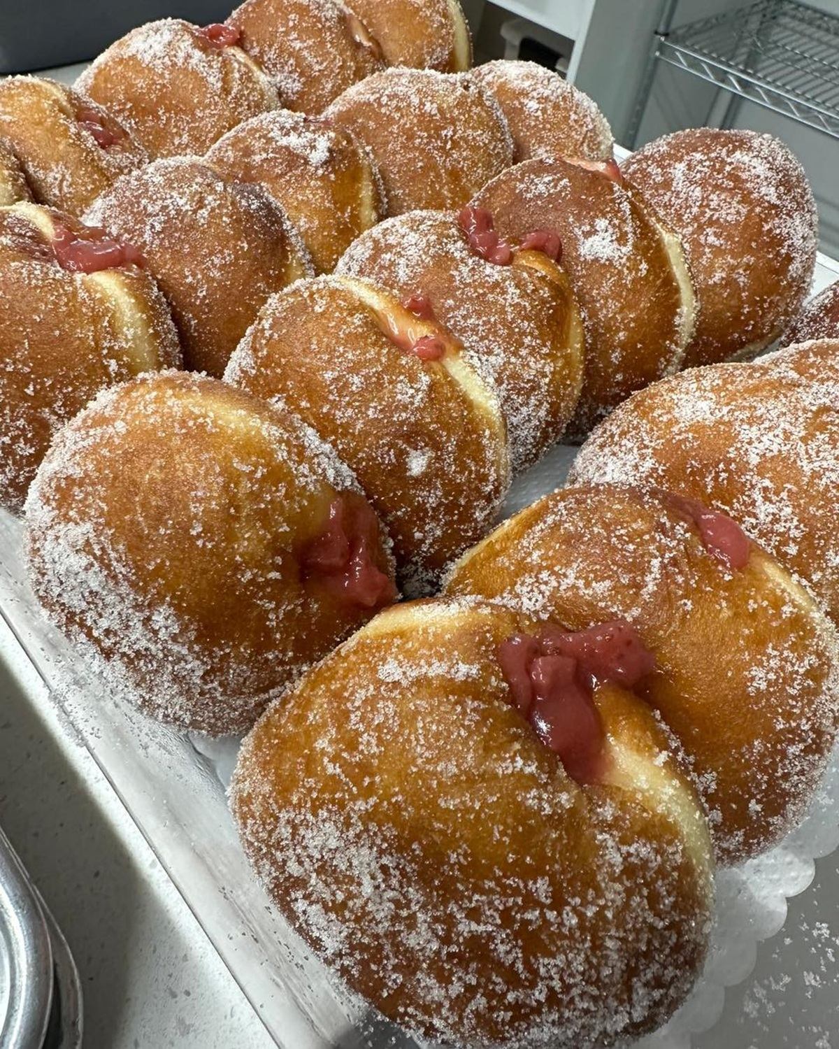 Fresh beignets dusted with icing sugar and filled with berry jam on a metal tray.