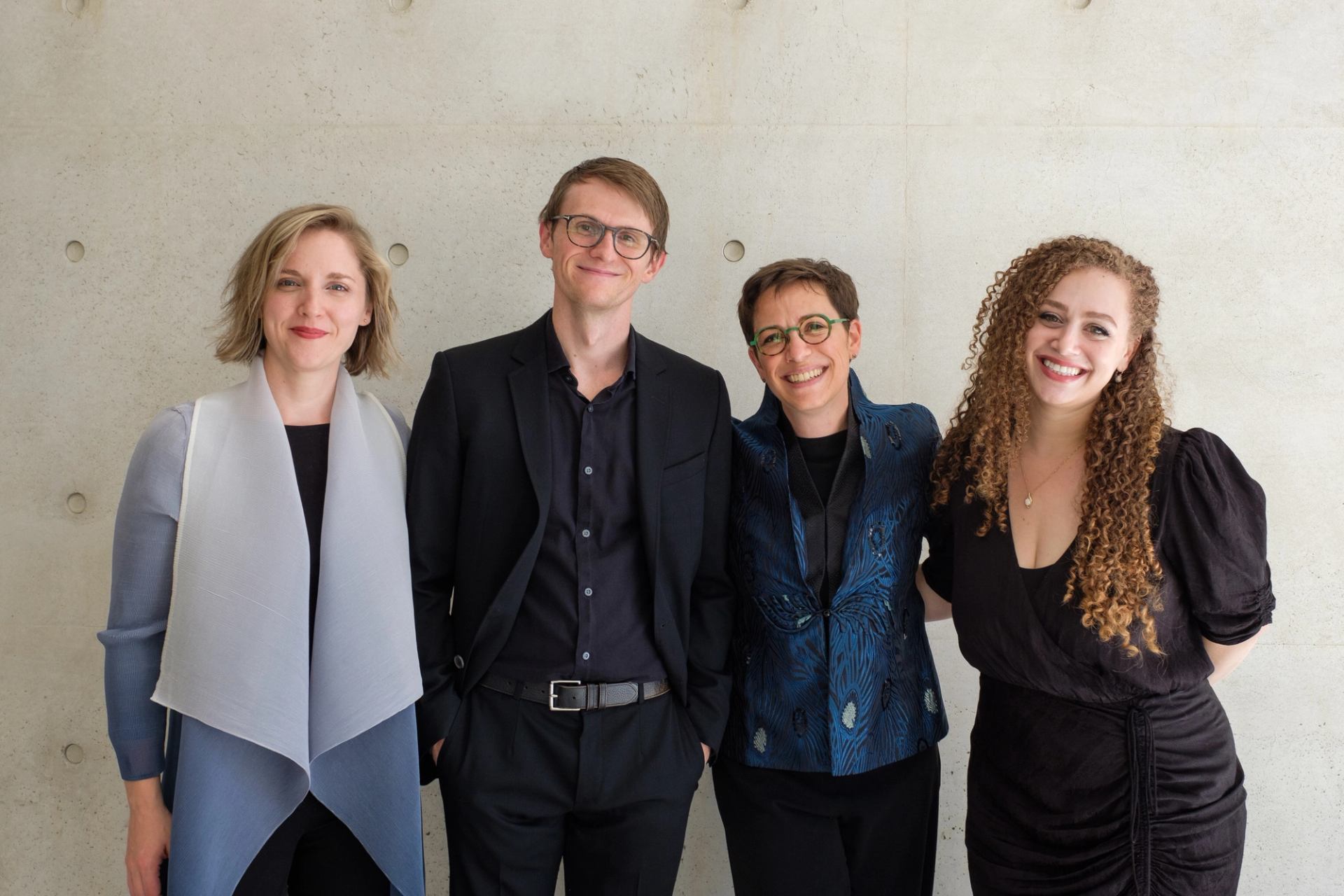 Four people stand side by side against a light concrete wall, facing the camera.