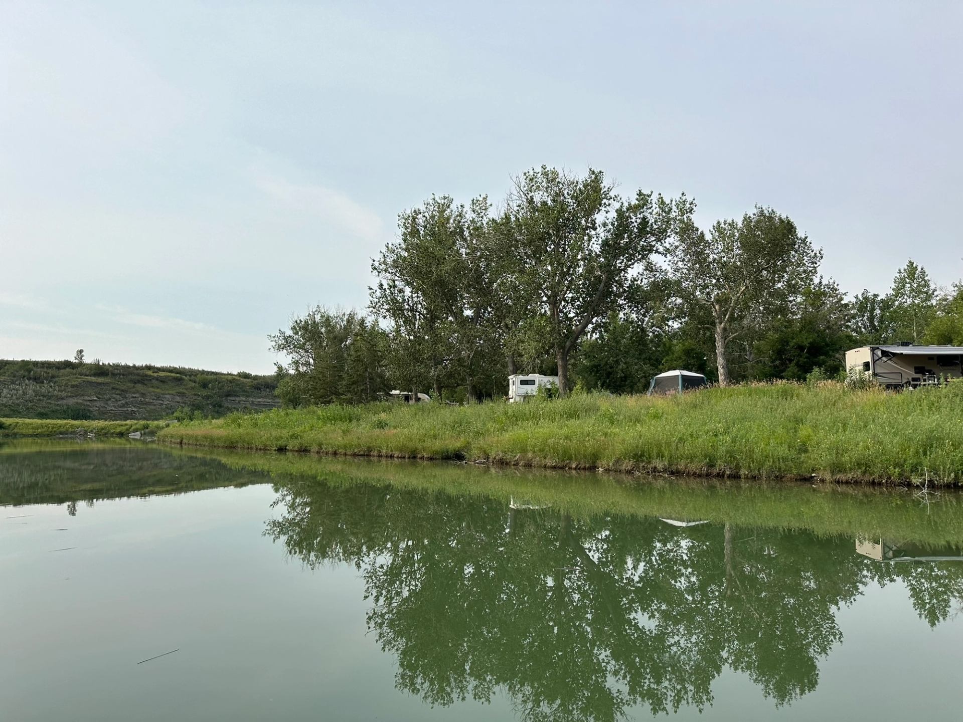 A calm river reflecting trees and RVs along a grassy bank.