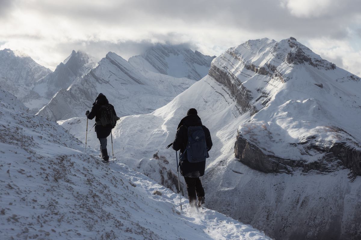 Two hikers trekking along a snowy ridge with towering winter mountains in the background.