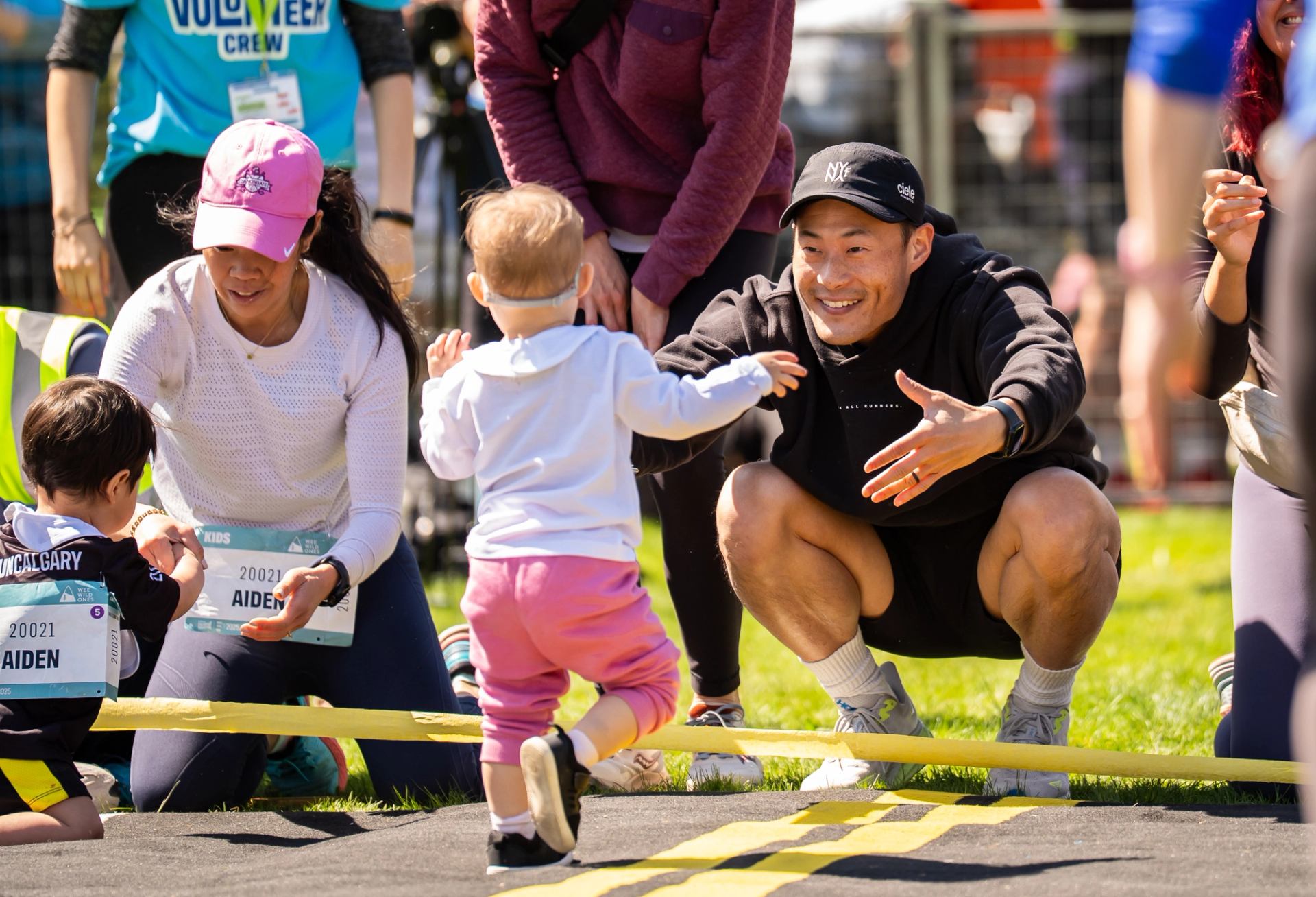 Adults crouch near course as toddler runs toward them by yellow tape.