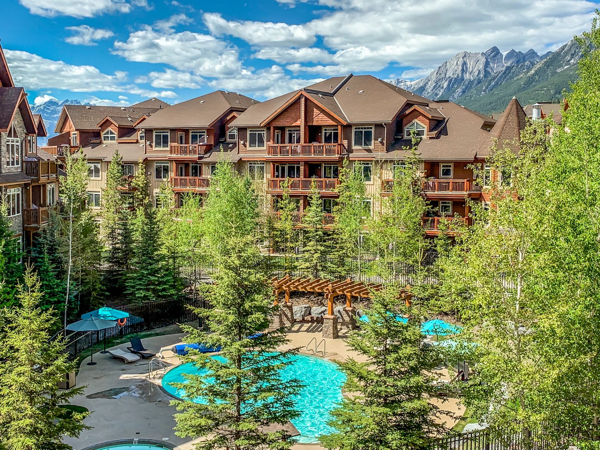 Mountain resort courtyard with outdoor pools, evergreen trees and multi-storey lodge buildings.