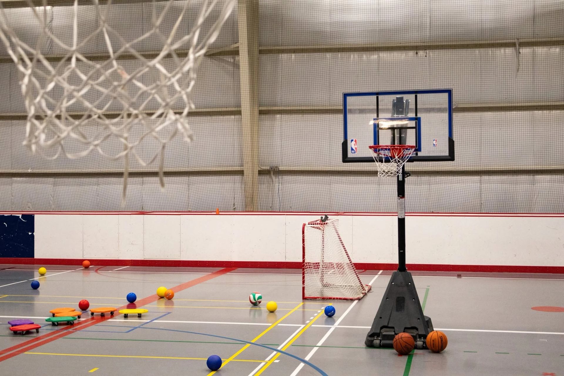 Indoor gymnasium with basketball hoop, floor scooters, balls and small hockey net.