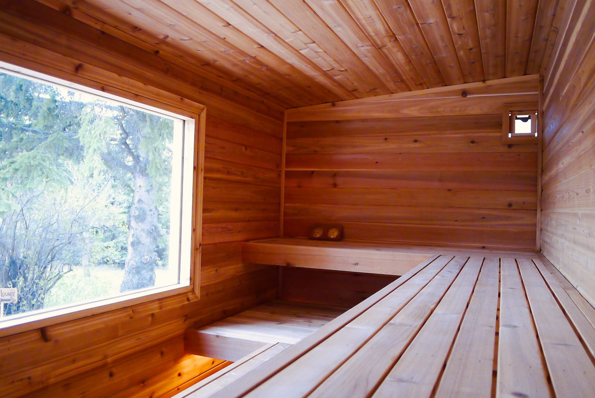 Inside a wooden sauna, smooth bench seating lines the walls with warm-toned wood panels throughout. A window looks out onto trees, bringing in natural light and views of the outdoors.
