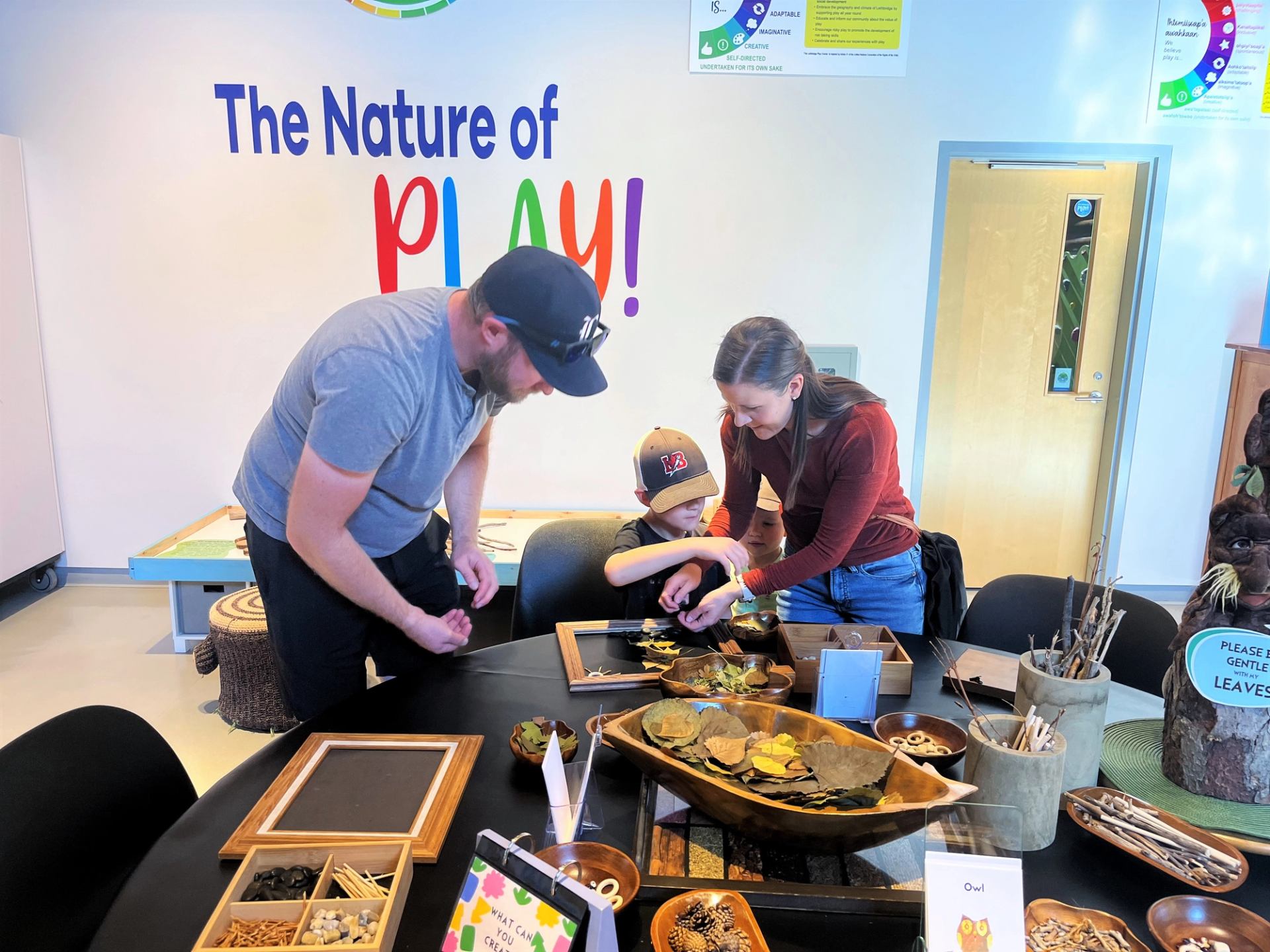 A man, woman, and young boy play with natural materials at a table in a bright indoor learning space.