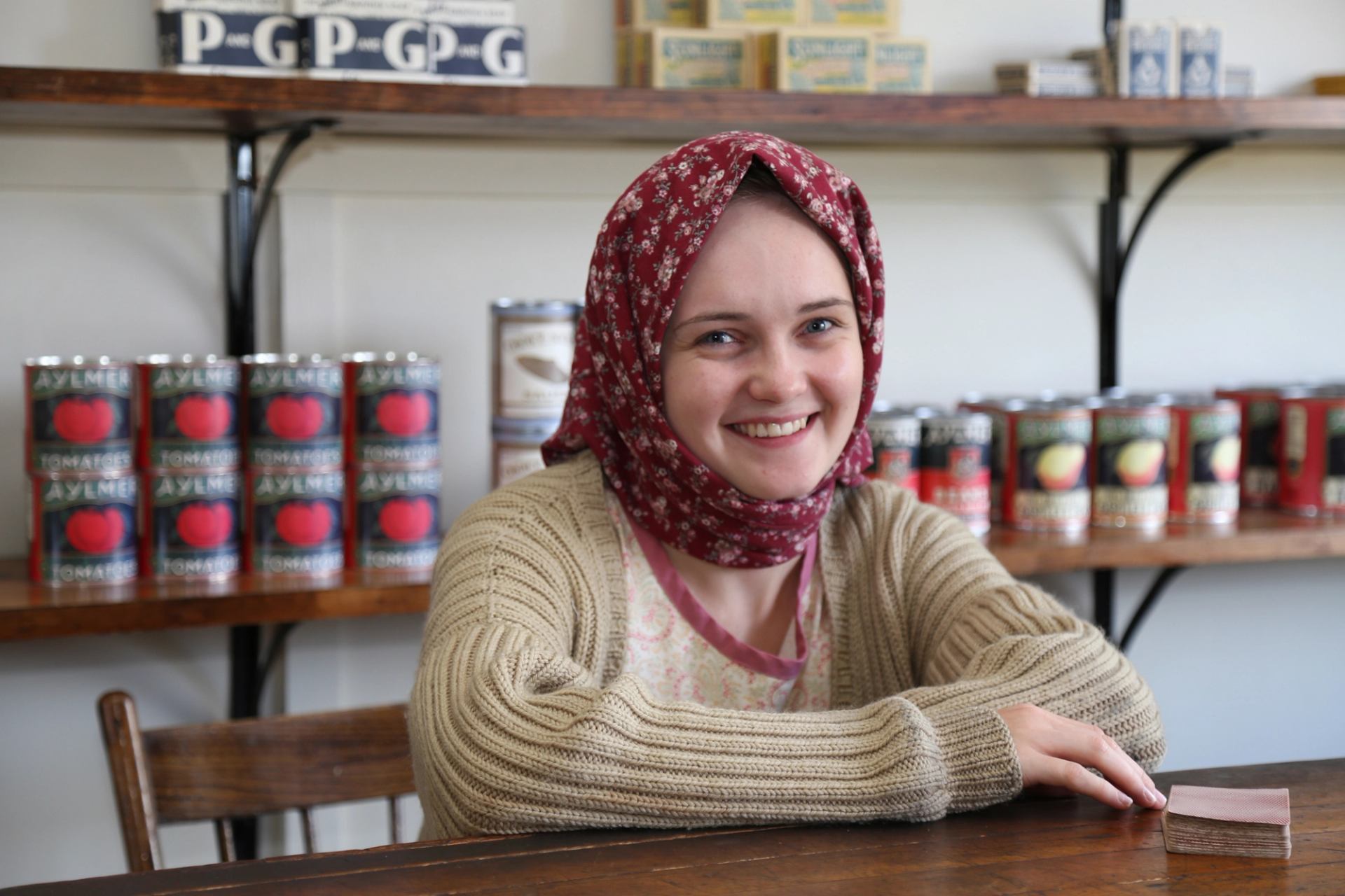 Person in floral headscarf sits at wooden counter with canned goods on shelves.