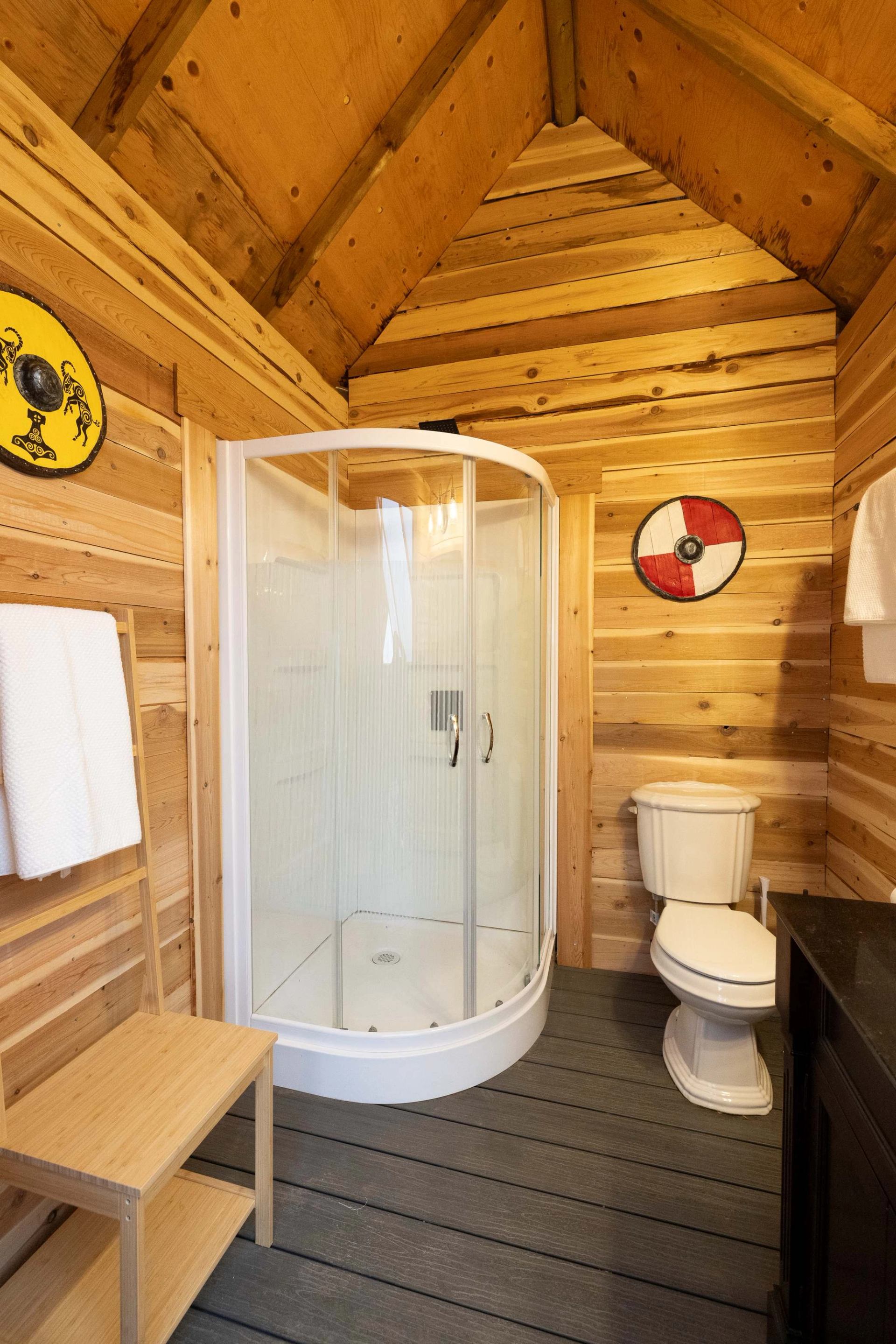 A rustic bathroom with wooden walls and ceiling, a curved glass shower, toilet, and two Viking-themed shields.