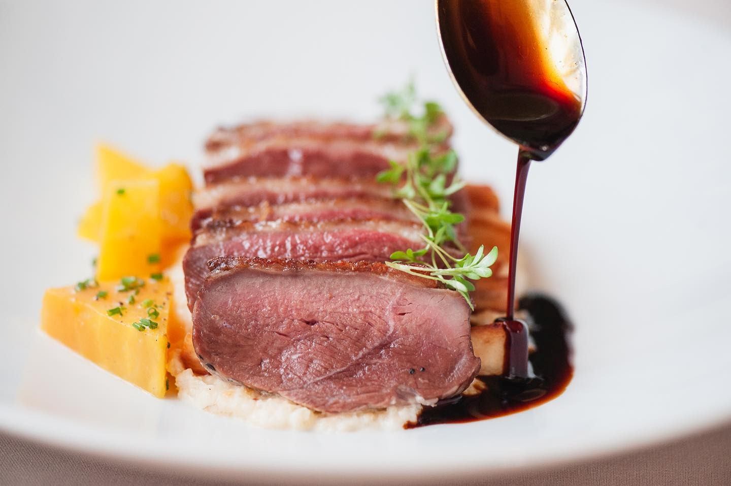 Sliced medium-rare duck on puree, garnished with pea shoots. A spoon drizzles dark sauce. Yellow cubes and herbs add contrast. Elegant presentation.