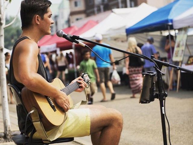 Musician playing guitar and singing at an outdoor market with colorful vendor tents.