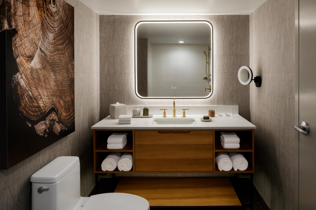 Bathroom vanity with backlit mirror, wood cabinetry, rolled towels and wall art.