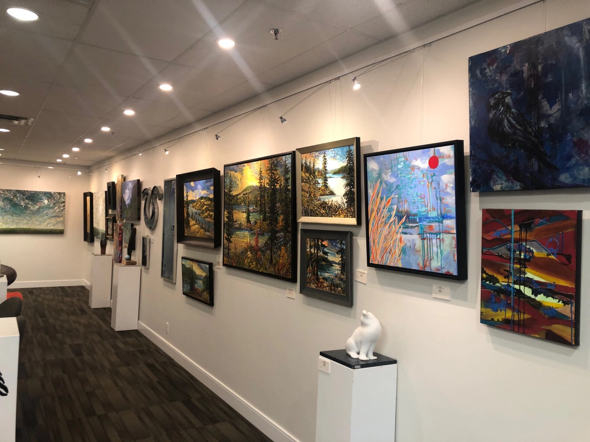 Art gallery display with framed paintings on walls and a white cat sculpture on a pedestal.