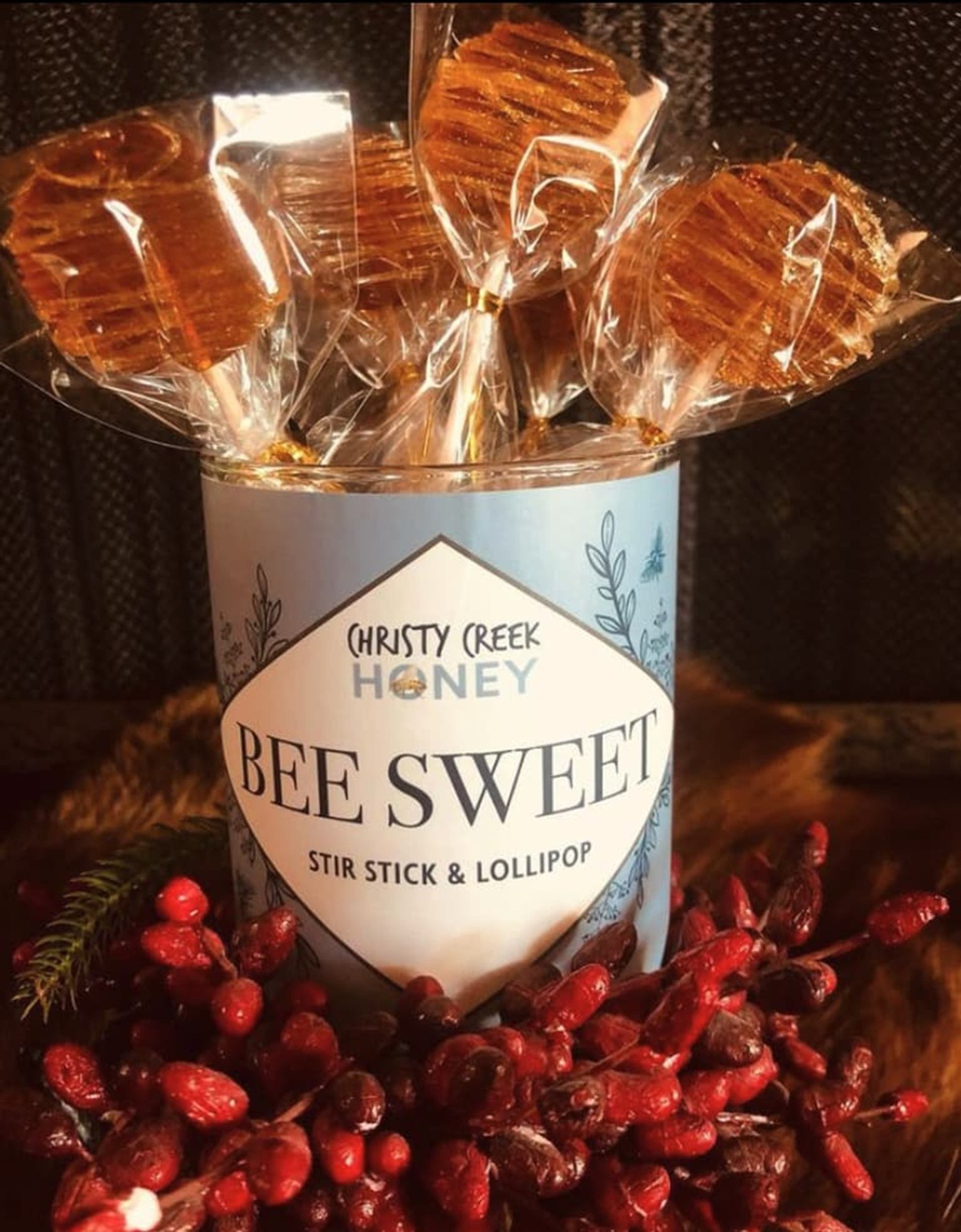 Bee Sweet honey stir stick lollipops in labelled tin, surrounded by red berries.