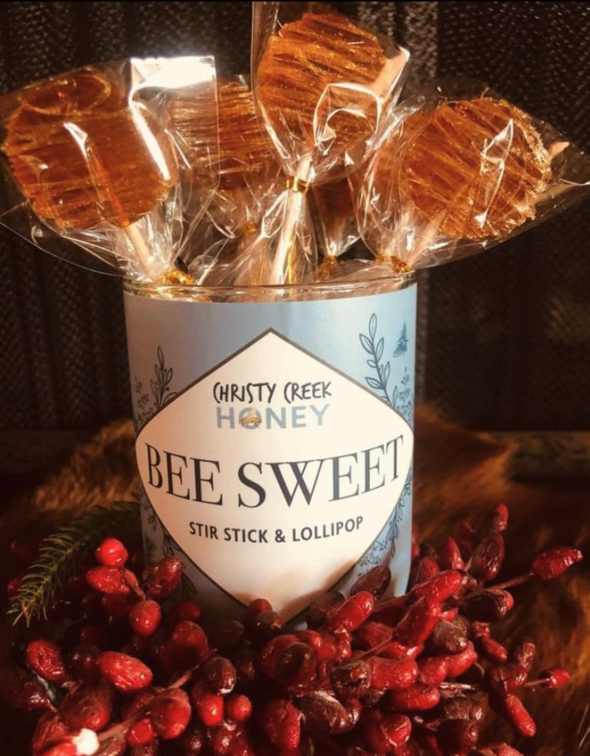 Bee Sweet honey stir stick lollipops in labelled tin, surrounded by red berries.