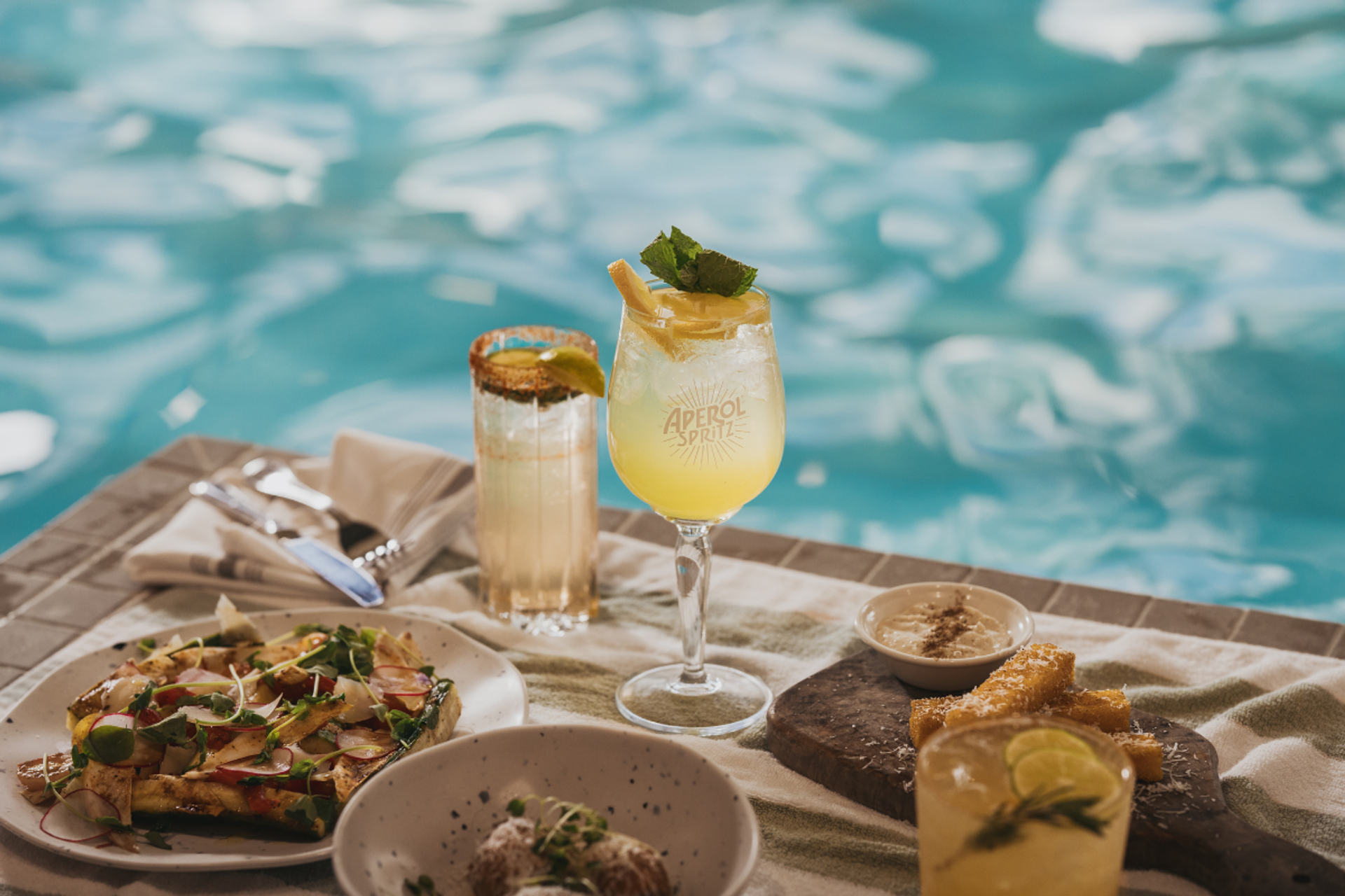 A vibrant spread of cocktails and appetizers, including an Aperol Spritz, laid out by a swimming pool.