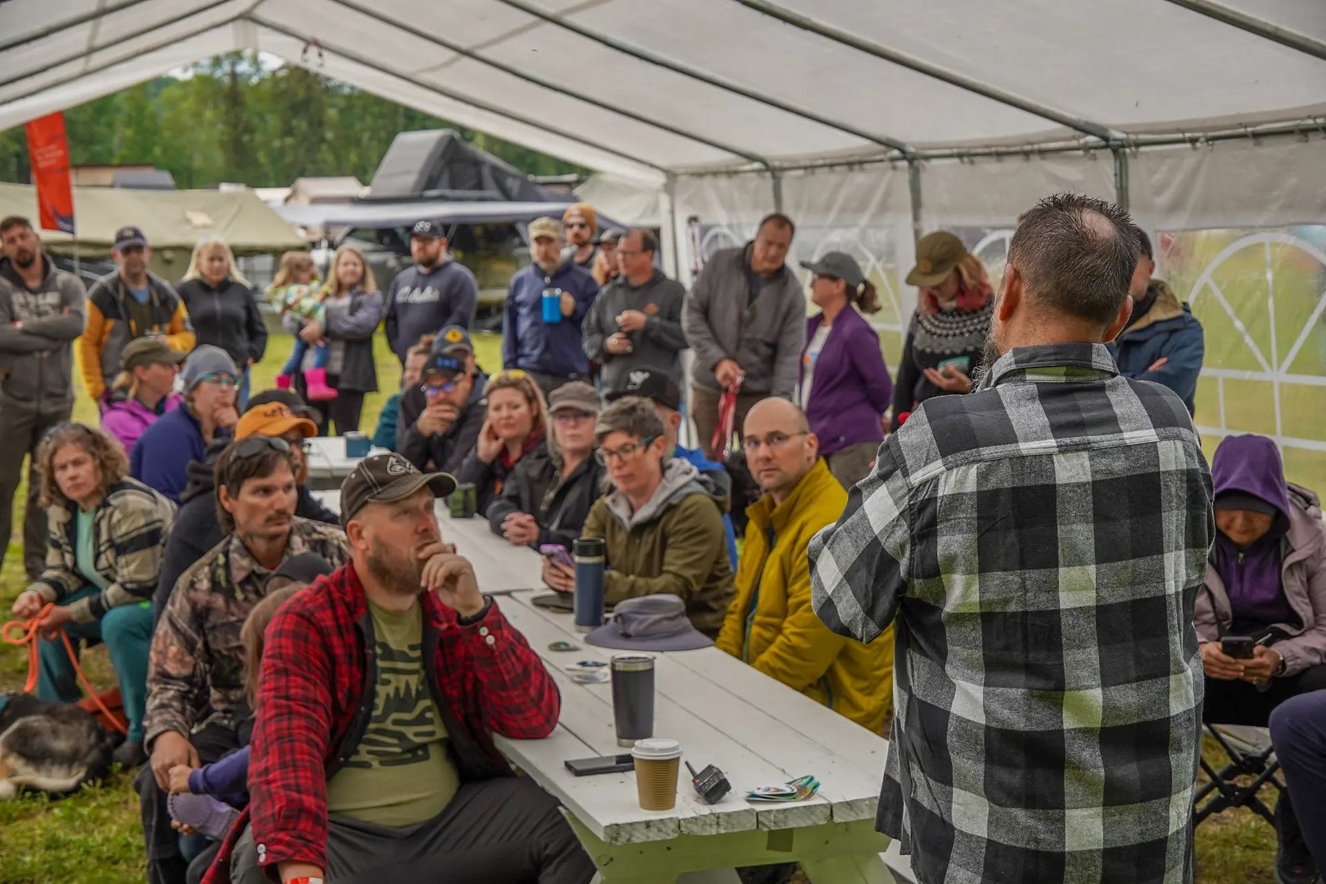 A diverse group of people seated and standing under a white tent, listening to a speaker at an outdoor event.