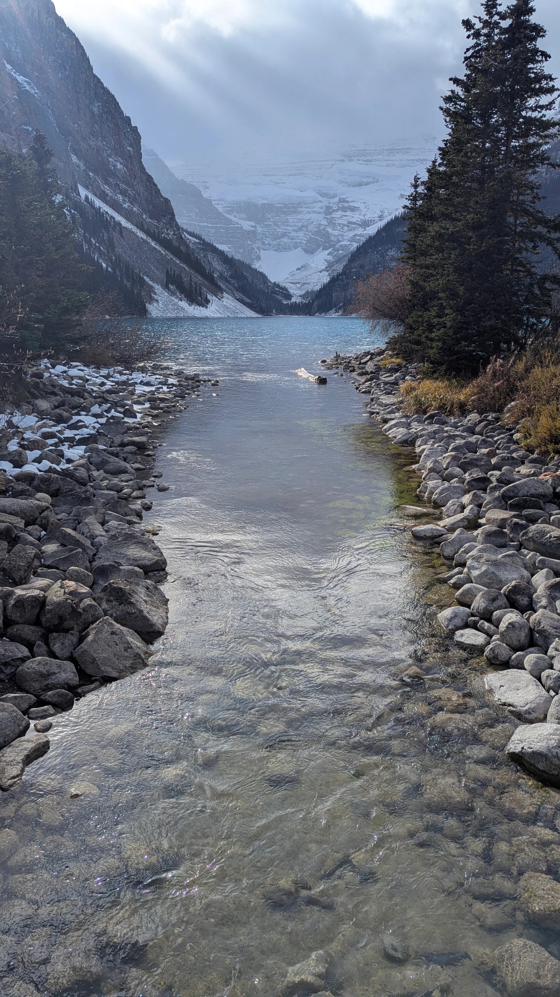 Clear stream flowing toward a turquoise lake with snowy peaks in the distance.
