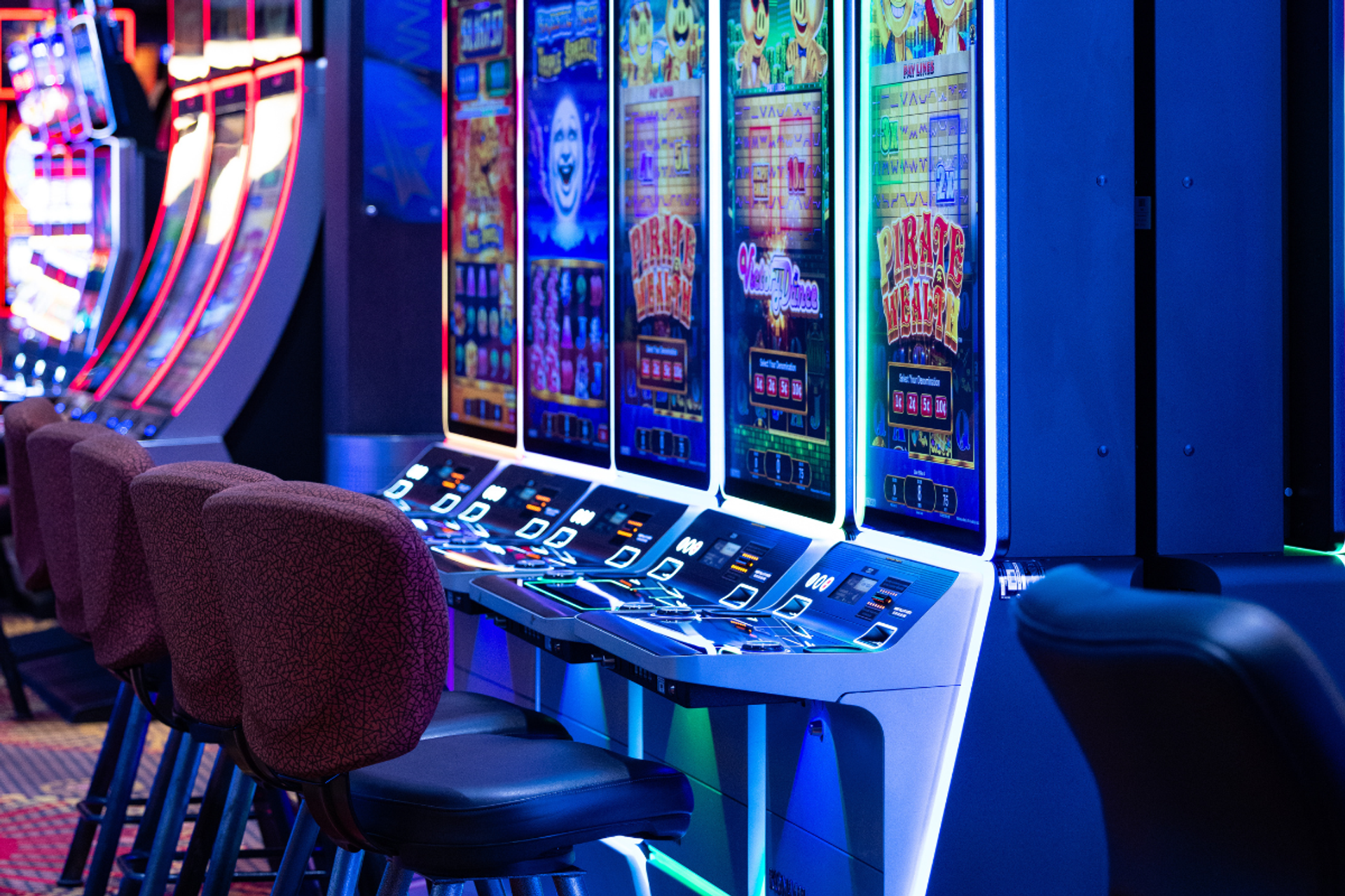 Row of illuminated slot machines with empty padded chairs in a dark casino interior.