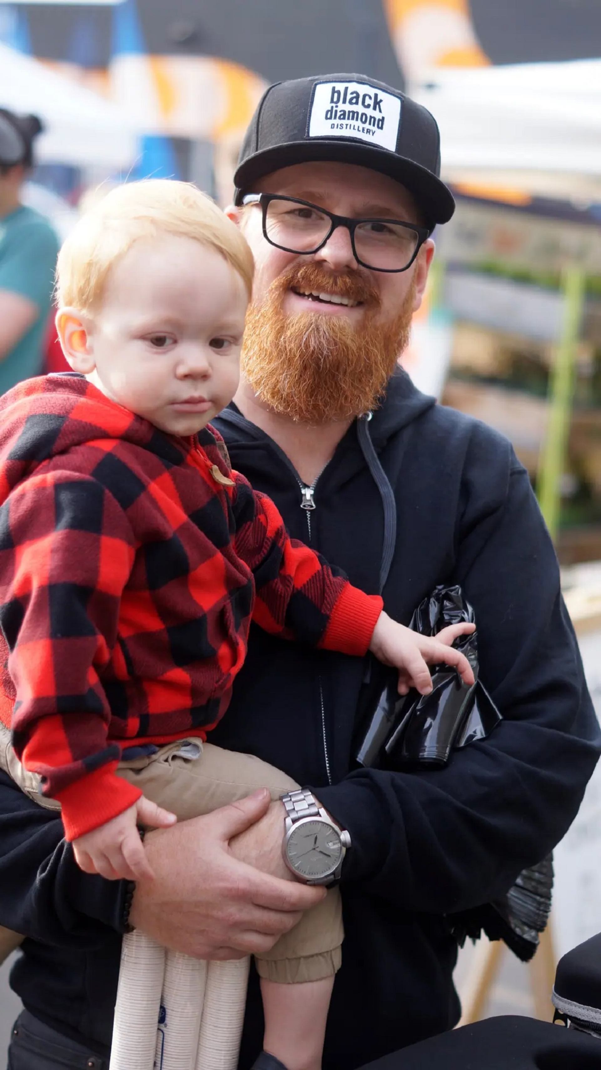 Adult holding a child while visiting an outdoor market with vendor tents in the background.